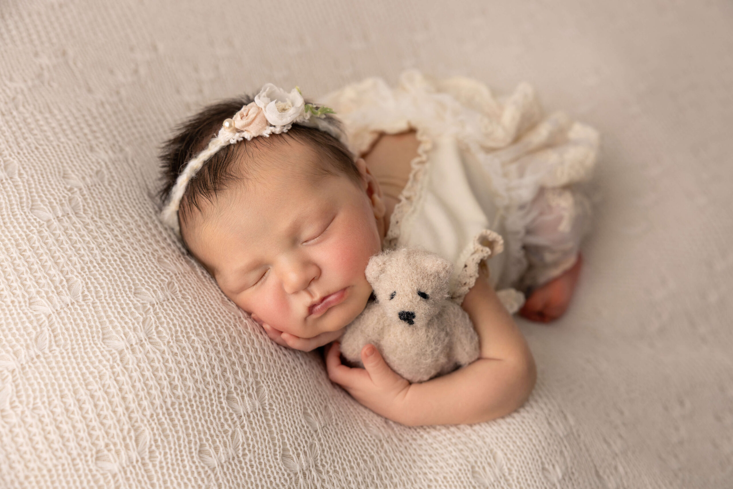 A newborn baby girl in a white lace dress cuddles a felt teddy in her sleep after visiting baby stores in Peoria, il