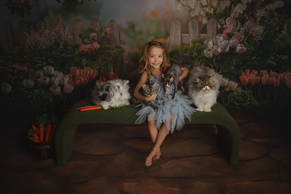 A happy toddler girl in a blue dress hugs two cats while sitting on a bench with two bunnies and another large fluffy cat