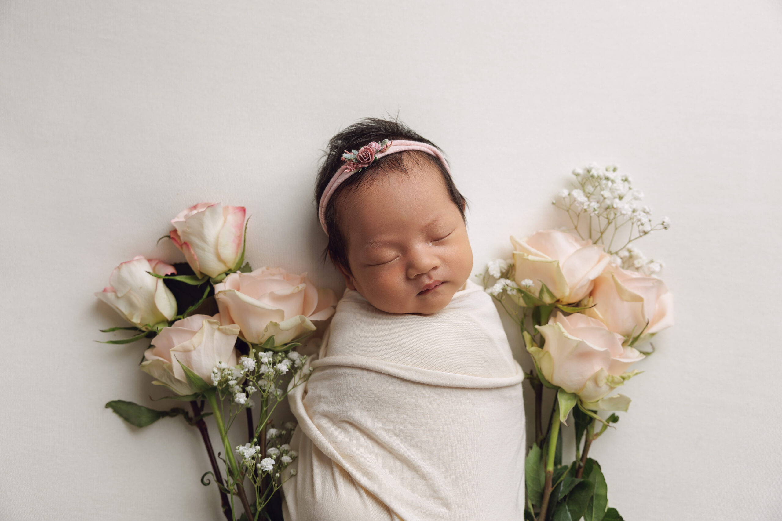 A sleeping newborn in a white swaddle surrounded by pink roses after finding pediatric dentists in peoria il