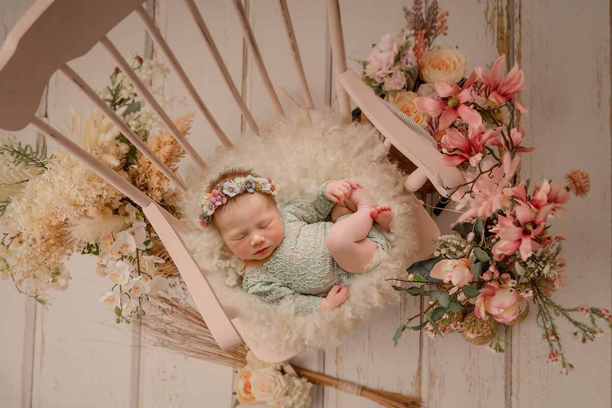 A newborn baby girl in a green lace onesie sleeps in a wooden rocking chair covered in pink flowers