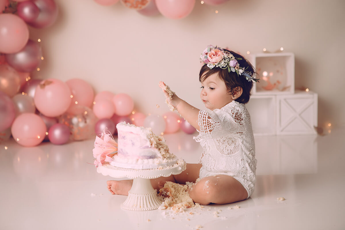 A happy toddler girl in a white lace onesie makes a big mess smashing her first birthday cake in a studio