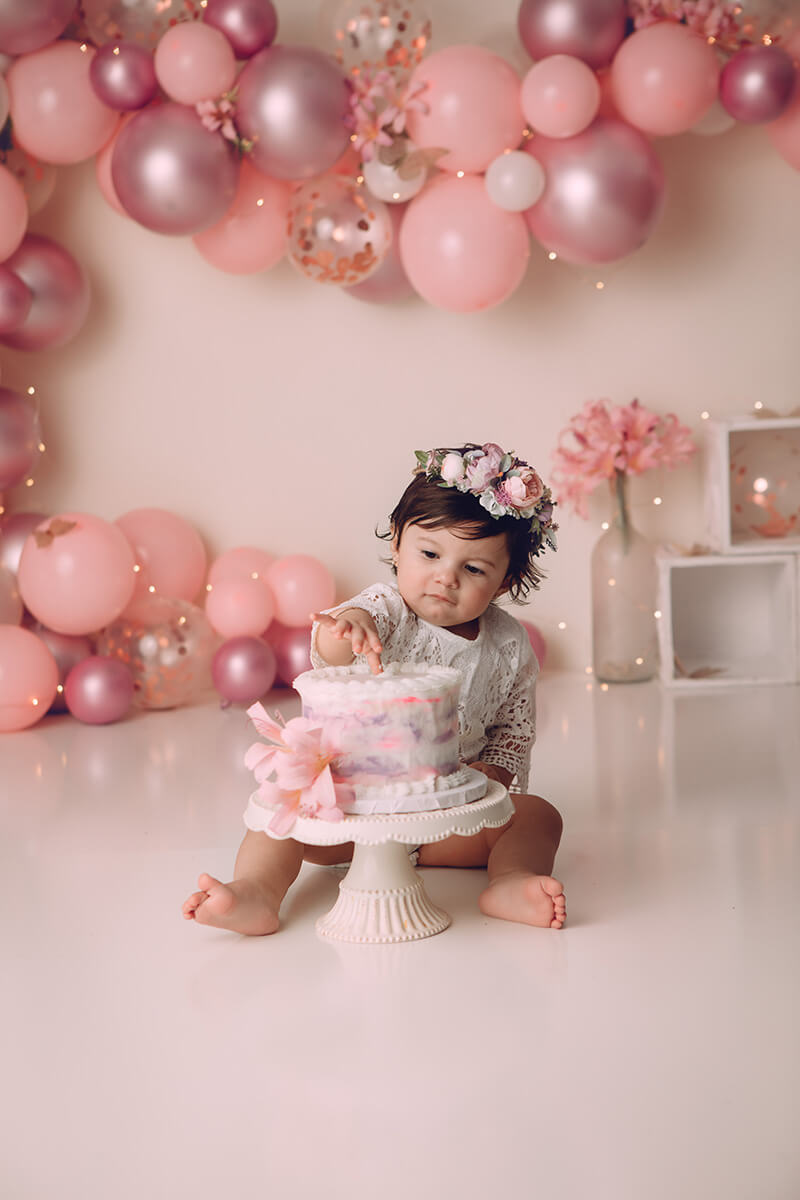 A toddler girl celebrates her birthday on the floor with a pink cake and a balloon wall after visiting Peoria Playhouse