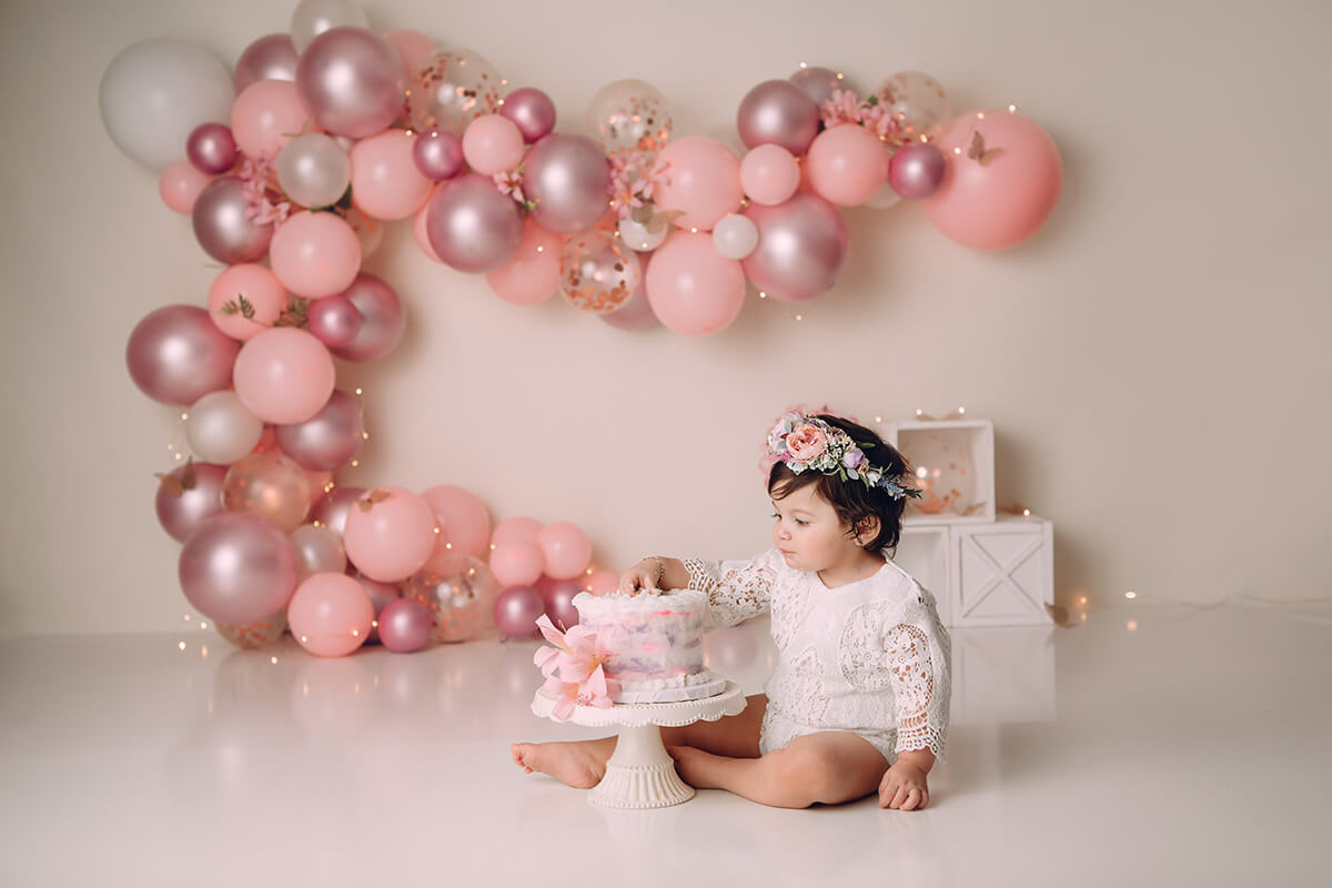 A toddler girl in white lace onesie and flower headband reaches in to taste cake on the floor of a studio after visiting Peoria Playhouse