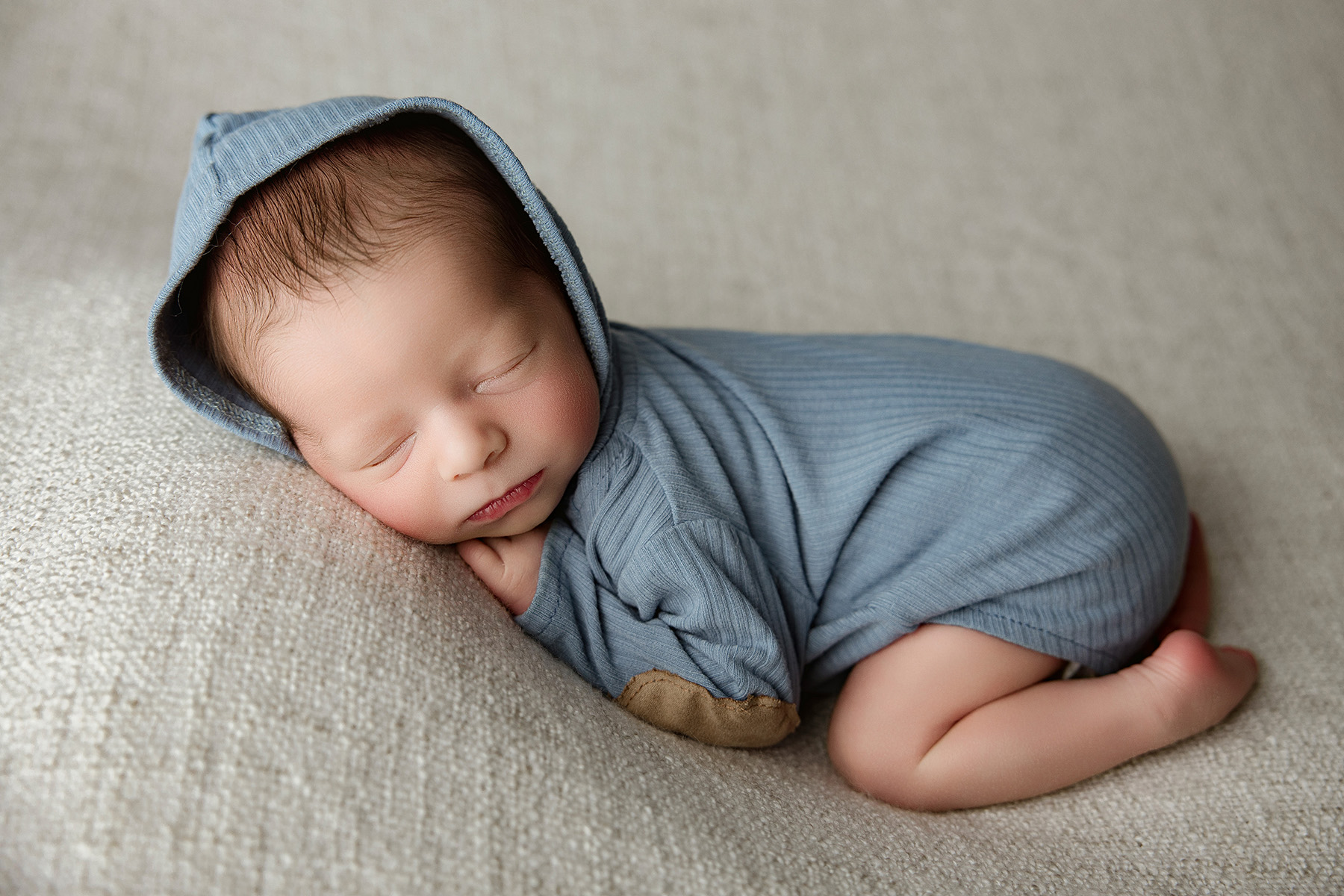 A baby boy sleeps in a blue onesie in froggy pose on a couch with a hood up after meeting lactation consultants in peoria il