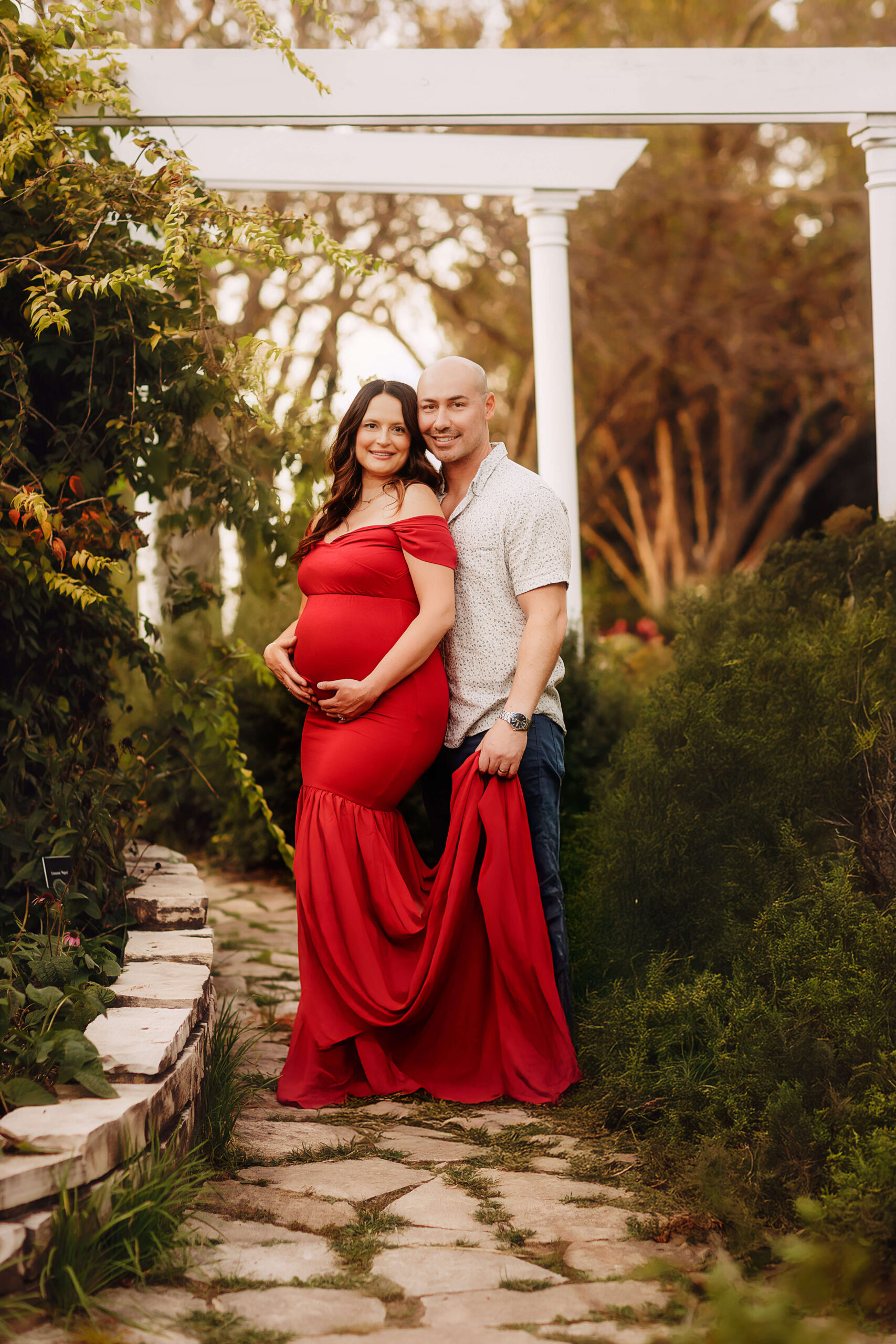 A pregnant woman stands leaning into the chest of her husband in a garden at sunset