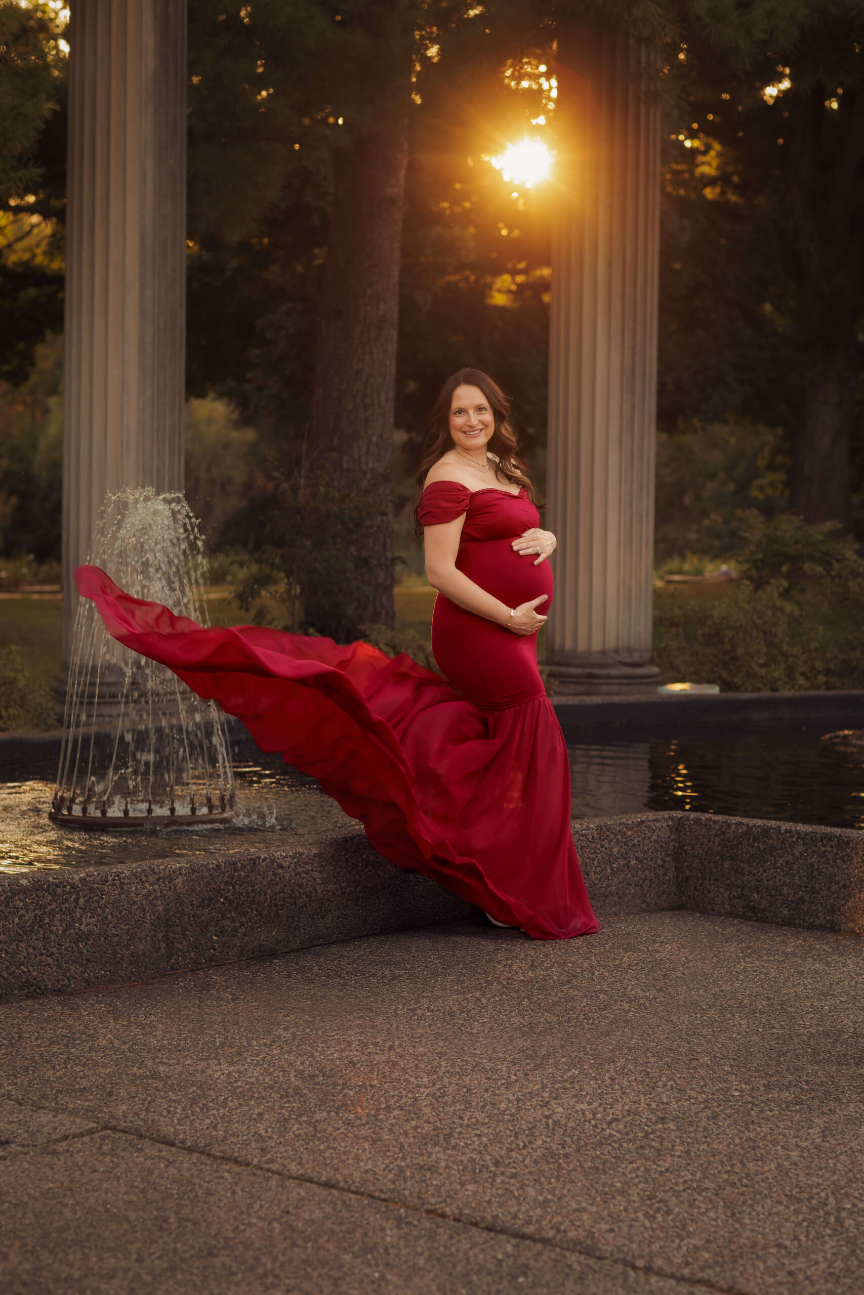 A smiling mother to be stands in an orate garden patio with a fountain and pond in a red maternity gown with train flying in the wind after finding a midwife in Peoria, IL
