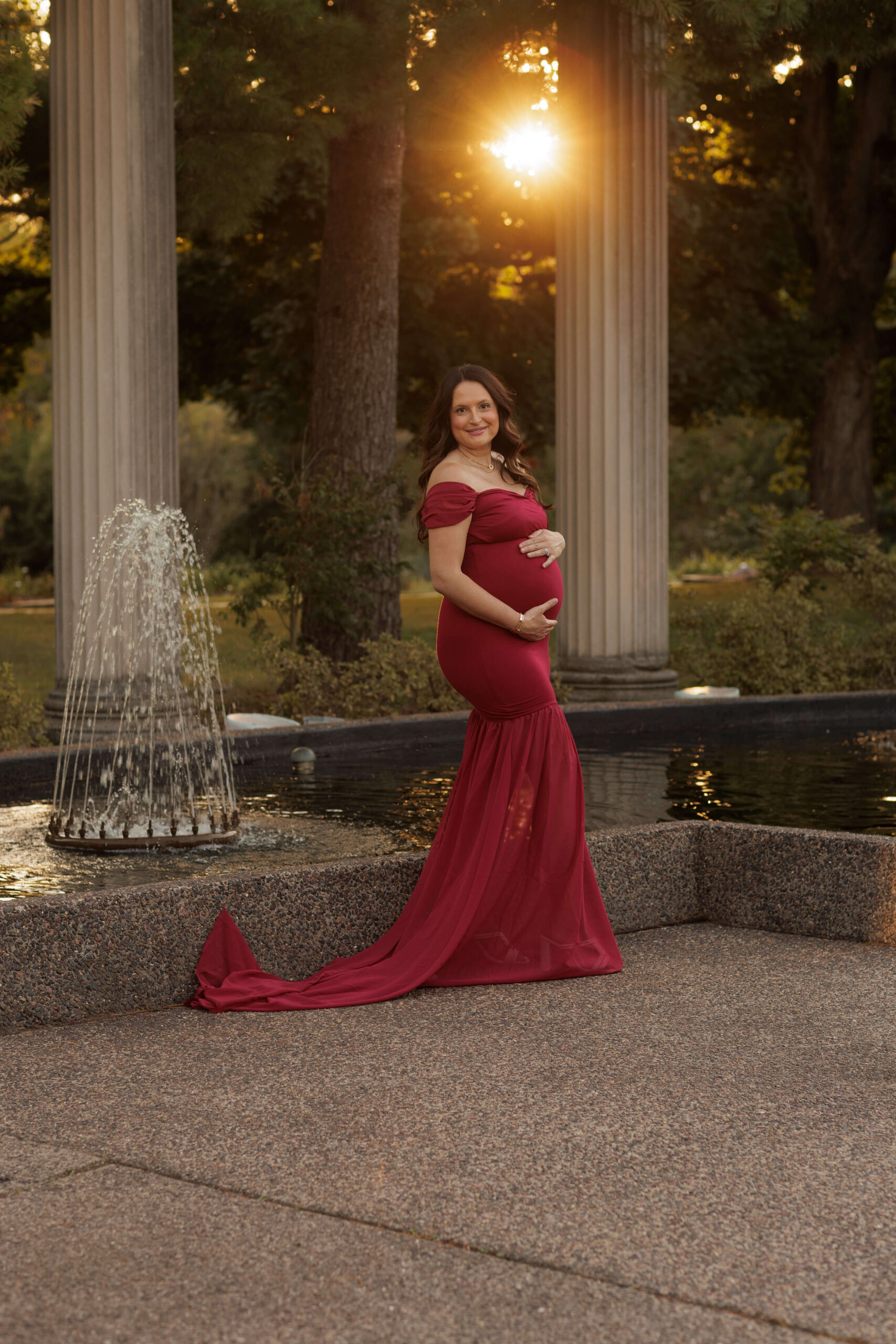A mother to be stands in a red maternity gown by a garden fountain after finding a midwife in Peoria, IL