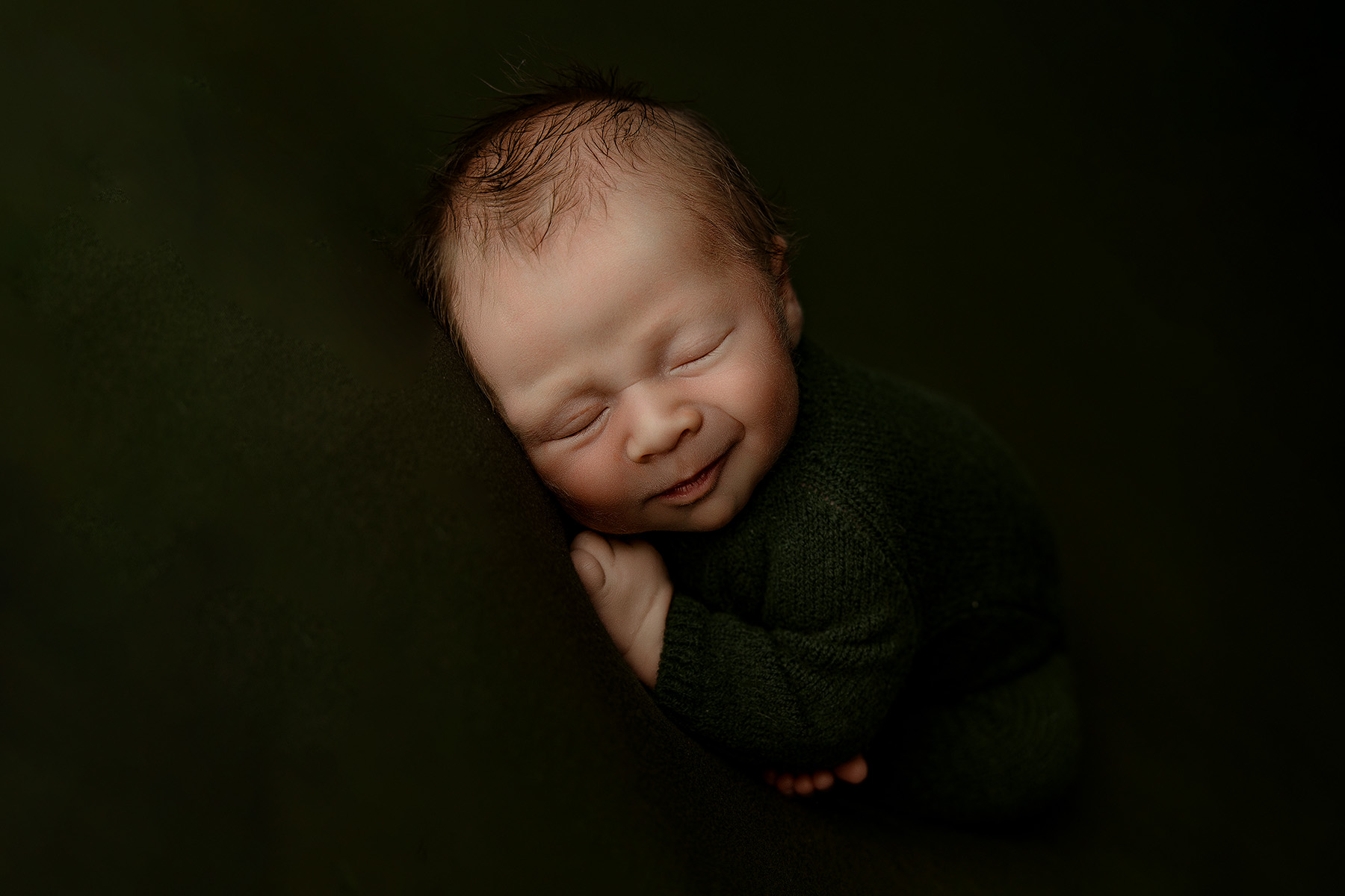 A newborn baby boy sleeps on his tummy in a dark studio in green