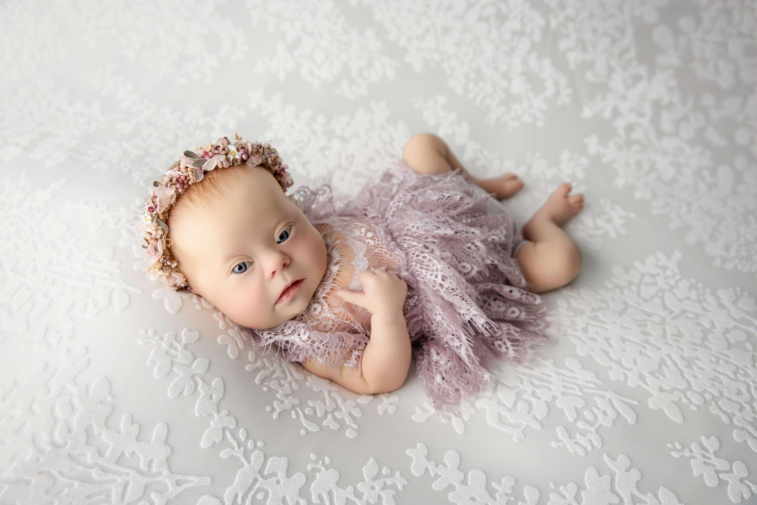 An awake newborn baby girl in a purple lace dress lays on a bed with eyes open and a flower headband