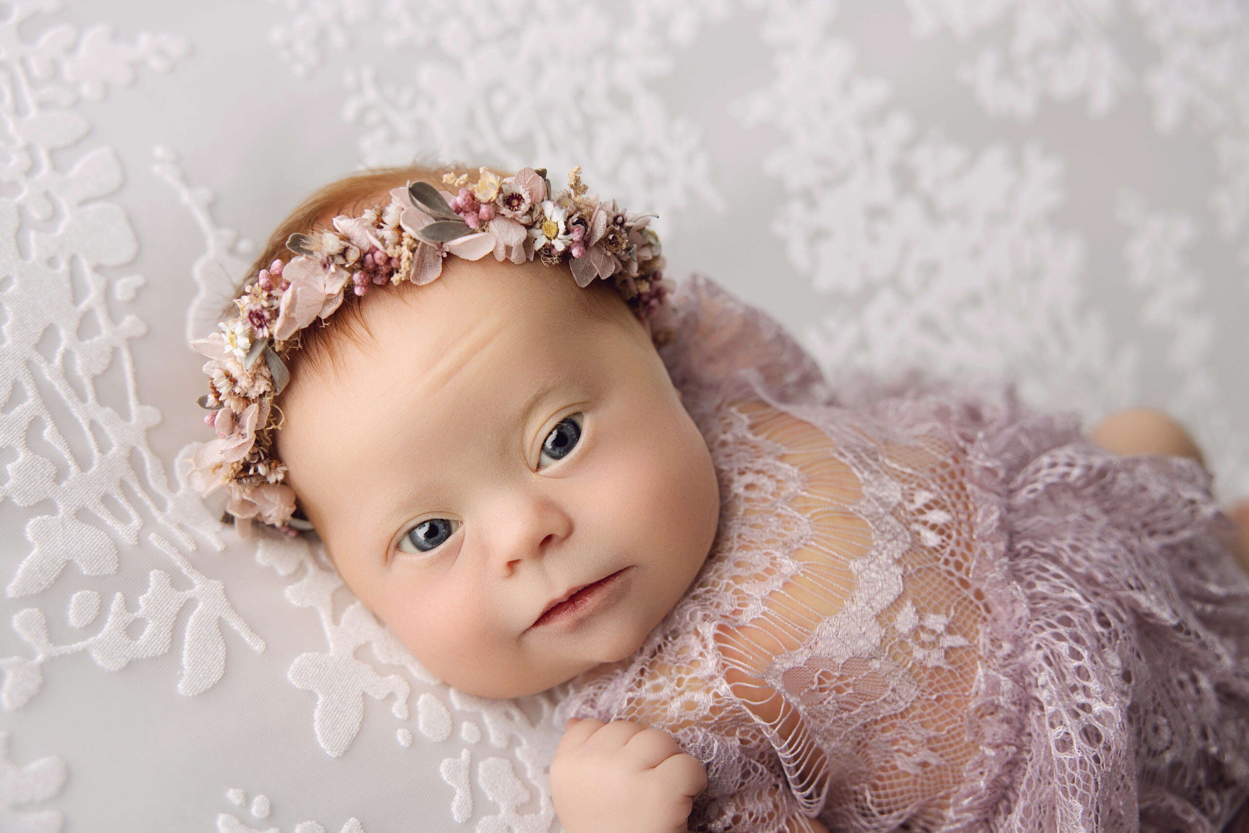 A newborn baby lays on a bed in a purple lace gown and flower headband with eyes open before exploring daycares in peoria il