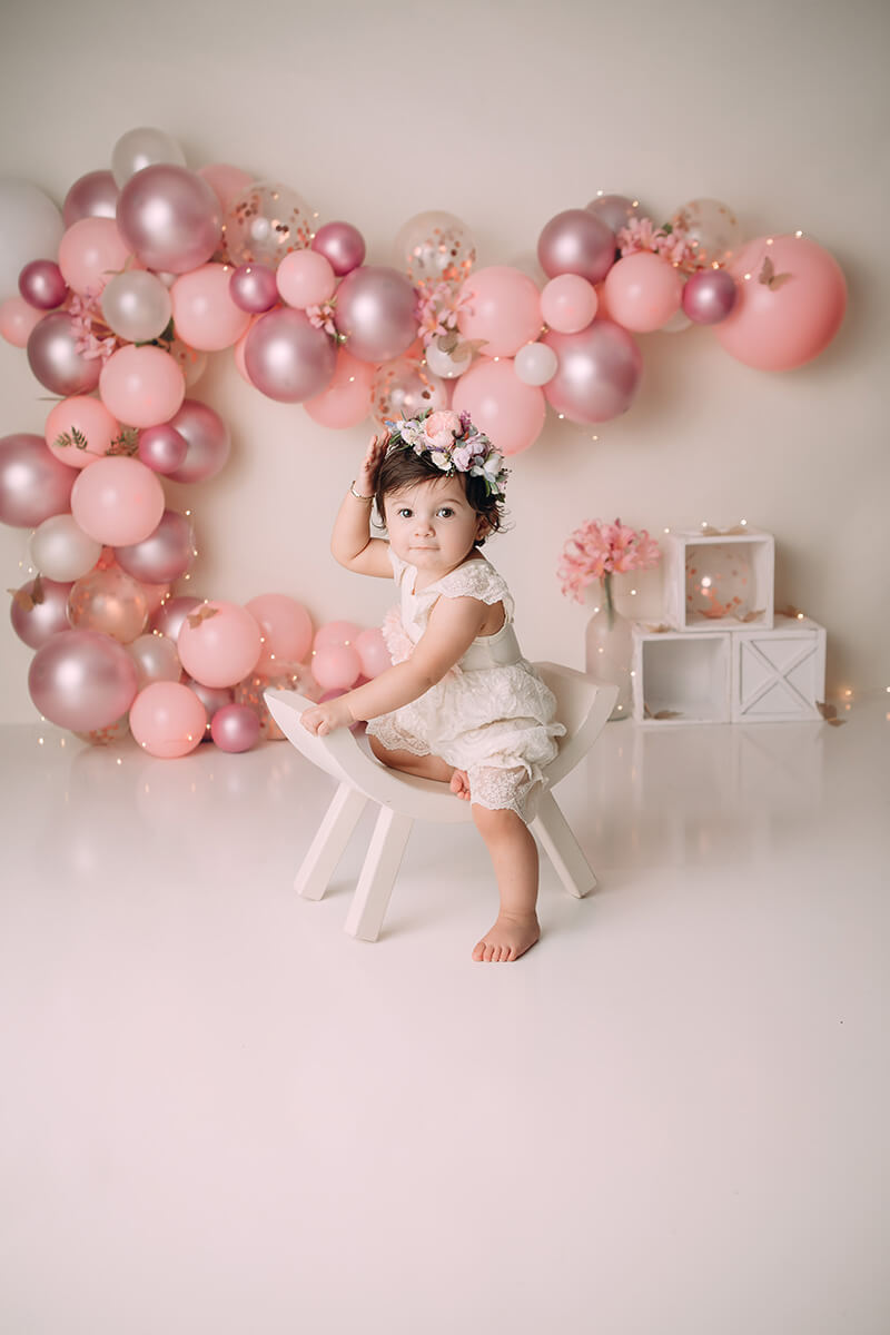 A happy toddler girl in a flower headband plays on a whtie curved stool in a studio