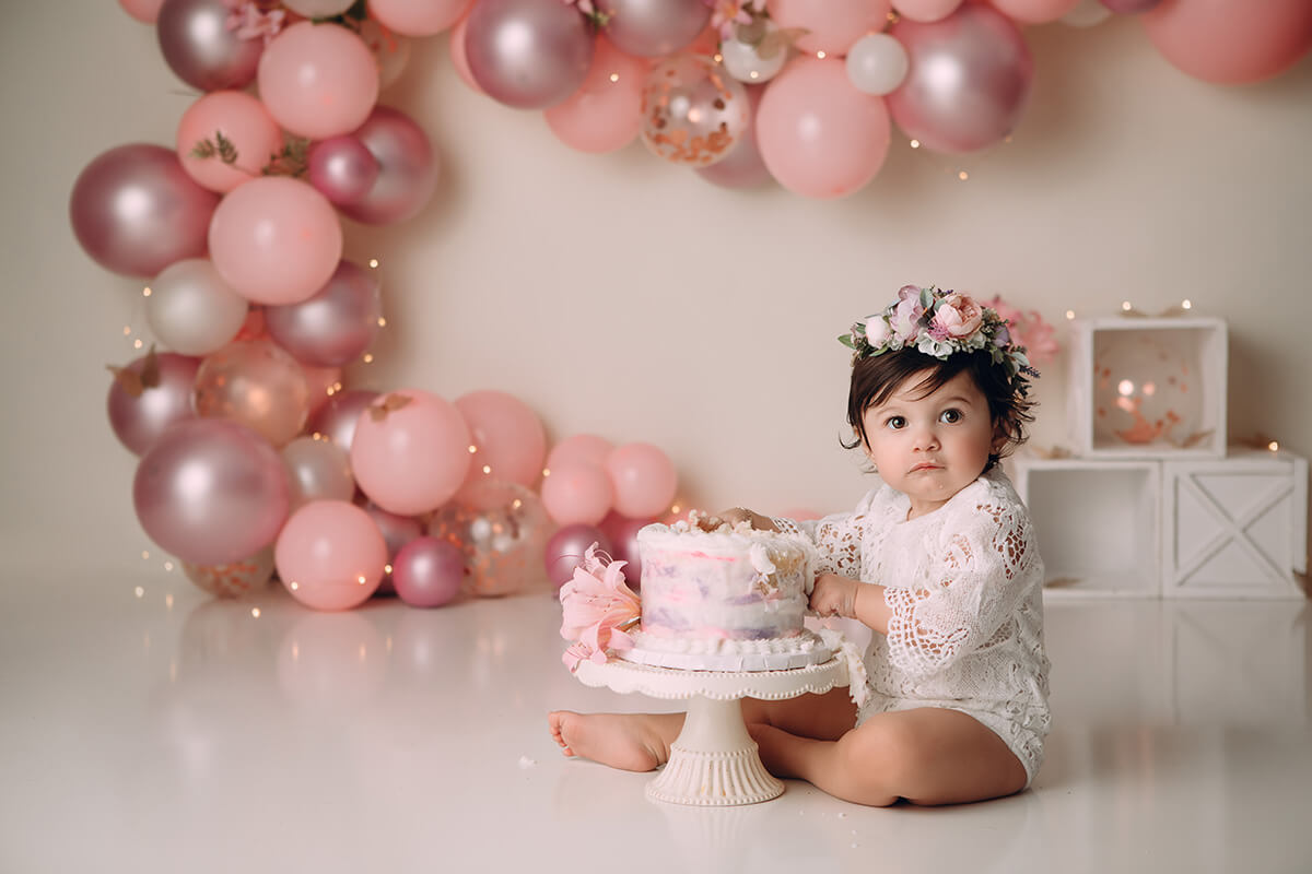 A toddler girl sits with her hands digging into a pink cake in a studio wearing lace
