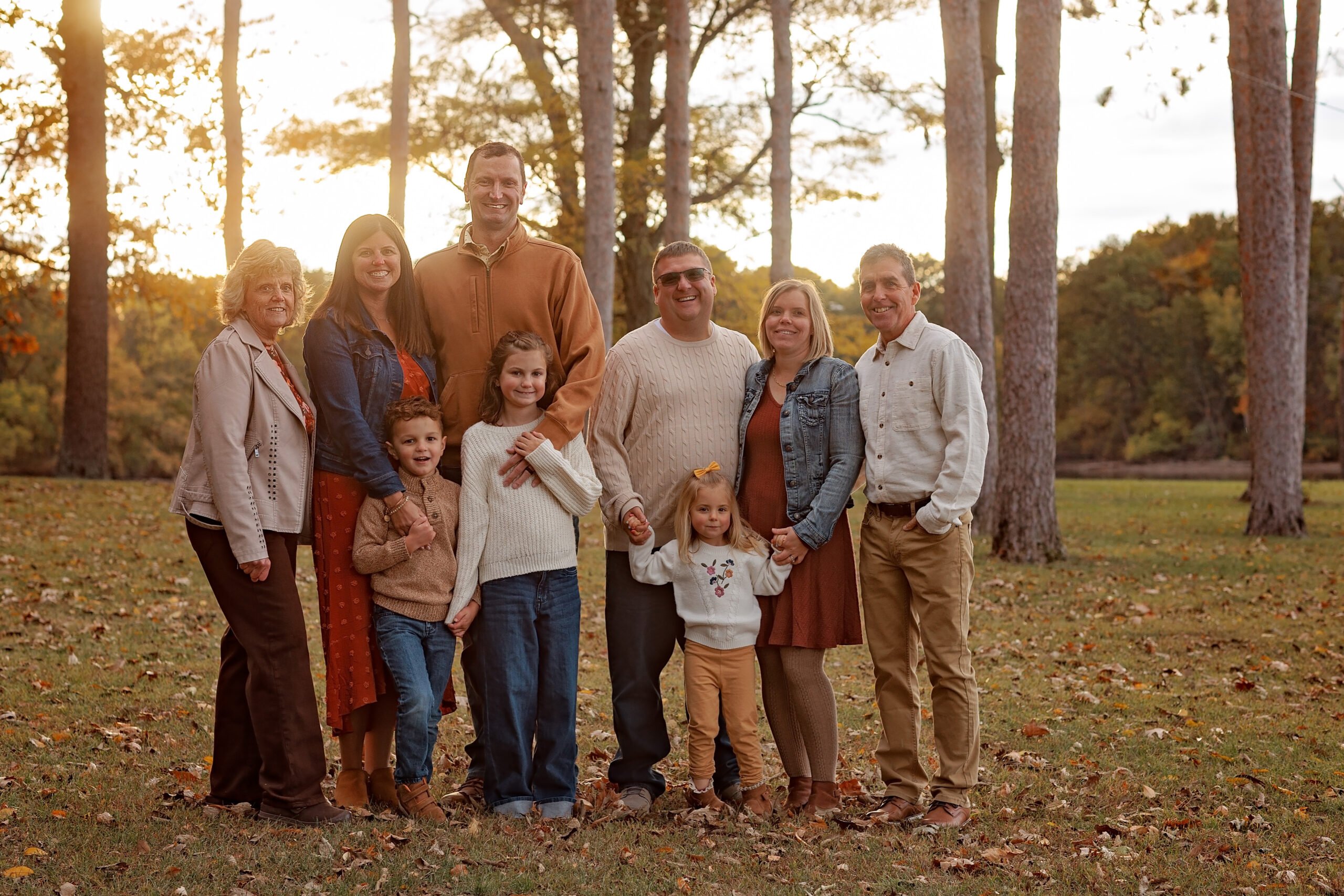 A smiling extended family in sweaters stands in a park lawn at sunset in fall