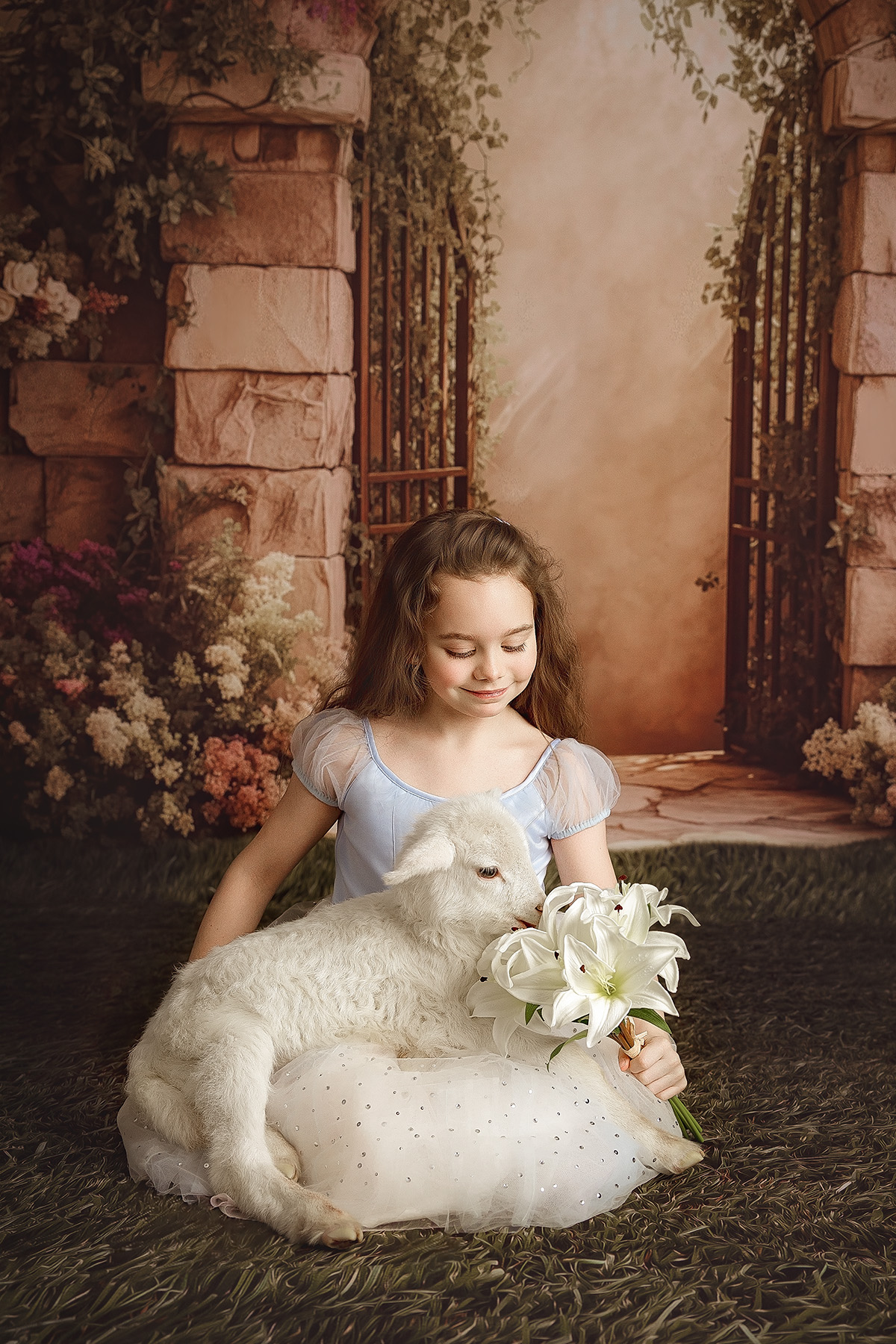 A smiling toddler girl sits with a lamb in her lap in a blue dress smelling flowers