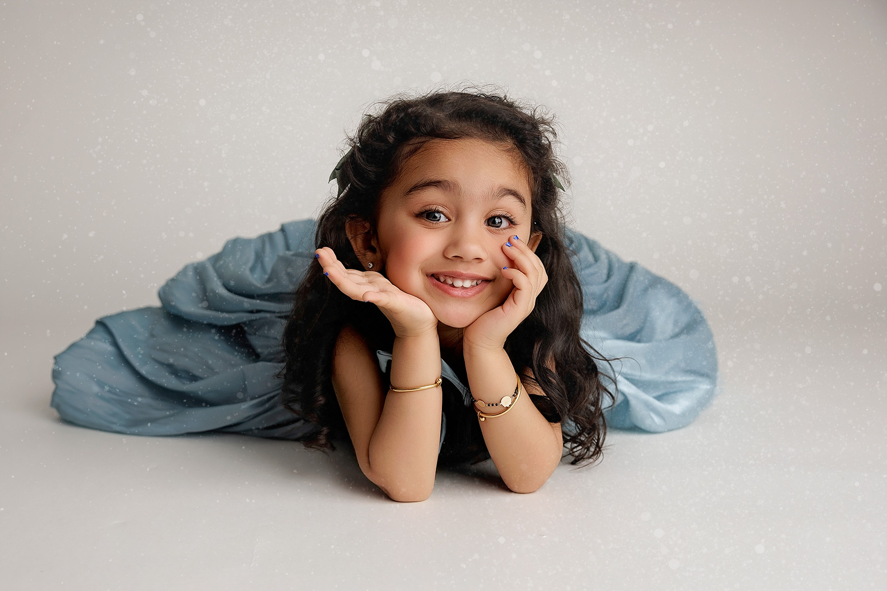A toddler girl in a blue dress lays smiling in a studio resting her chin on her hands after finding preschools in Peoria, IL