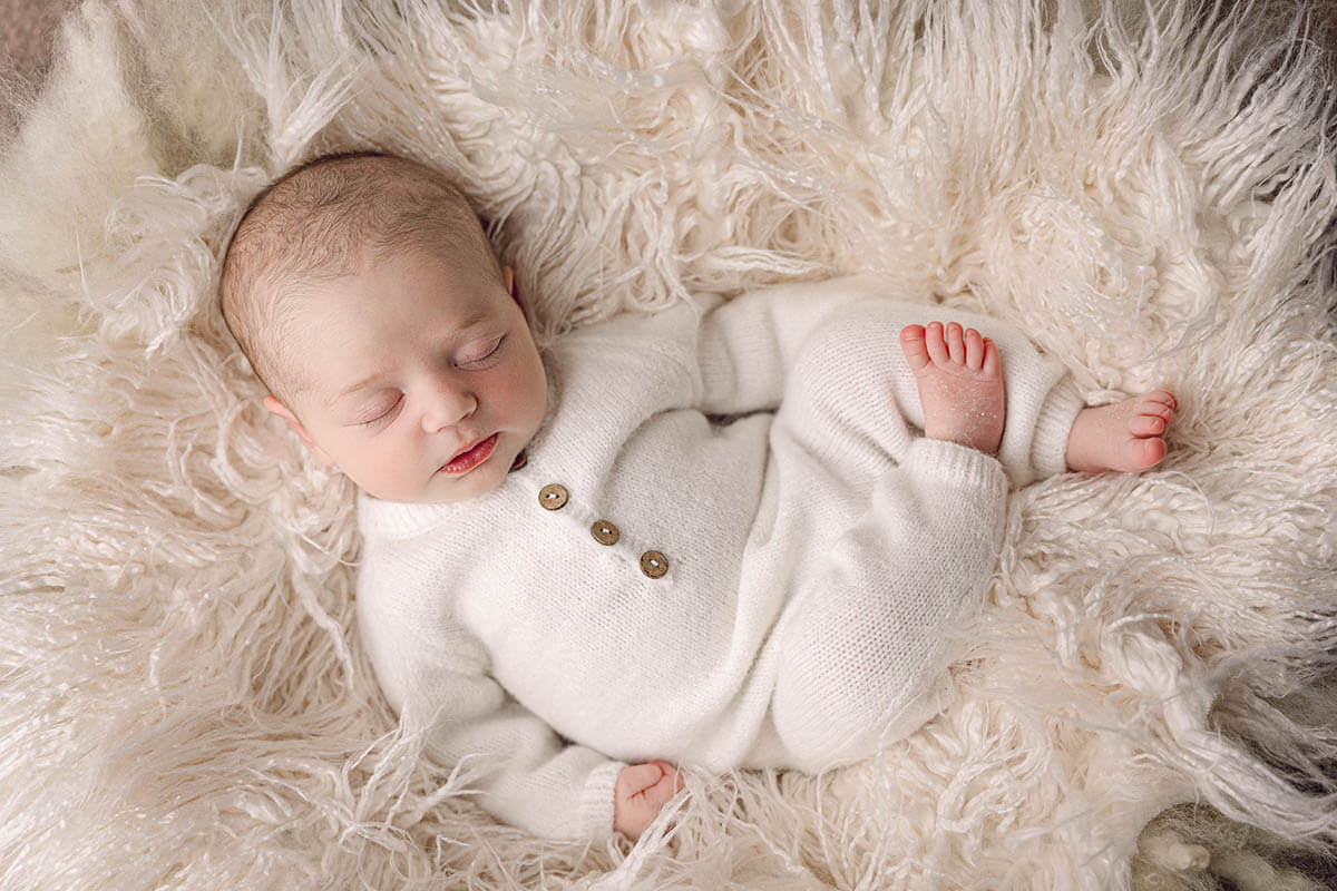A newborn baby sleeps in a white onesie on a fur blanket
