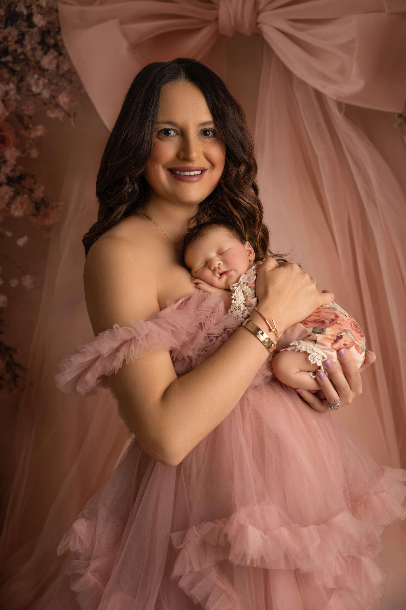 A woman in a pink tule maternity gown cradles her newborn girl sleeping against her chest in a studio