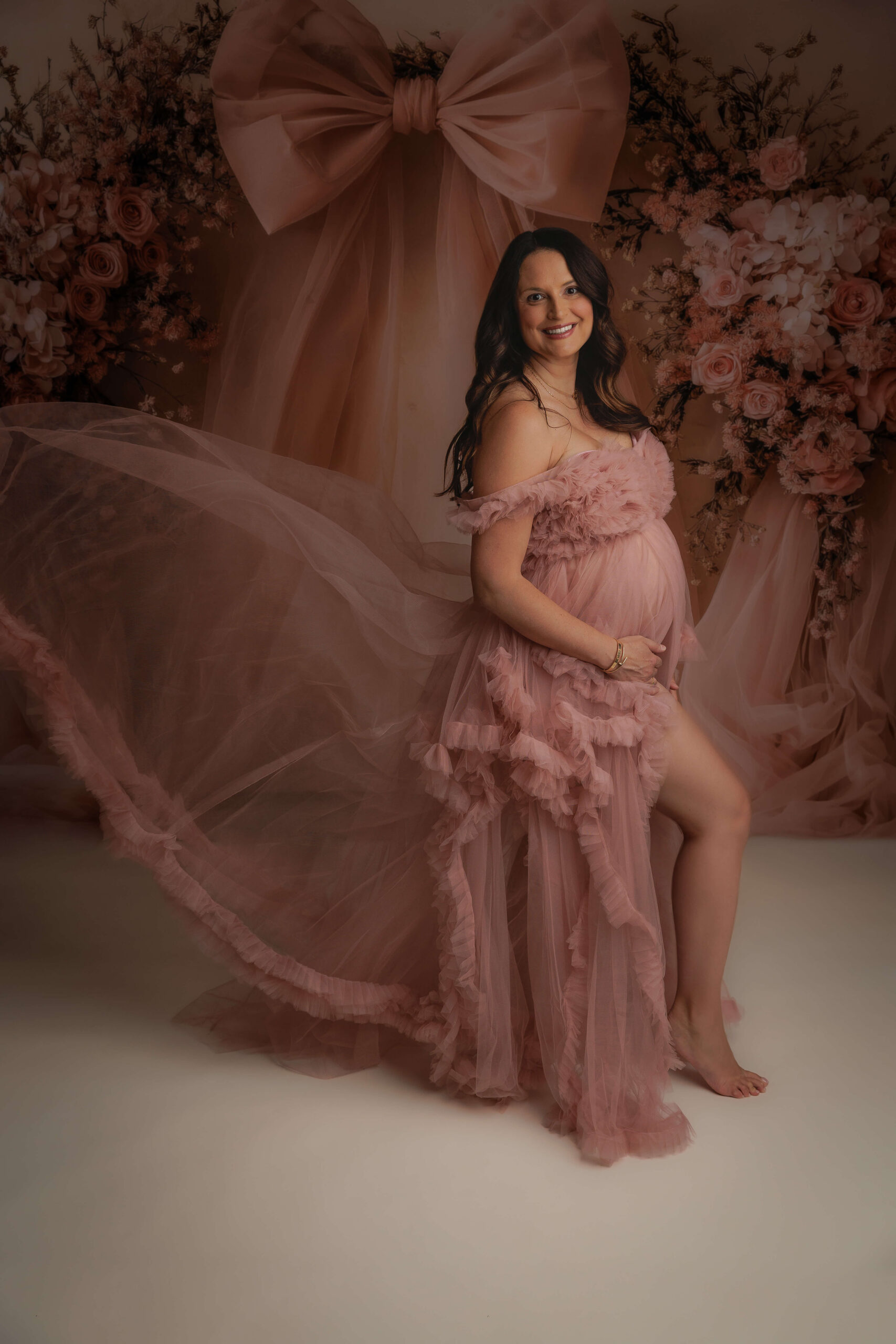 A mother to be in a pink tule maternity gown stands in a studio smiling after a prenatal massage in peoria il