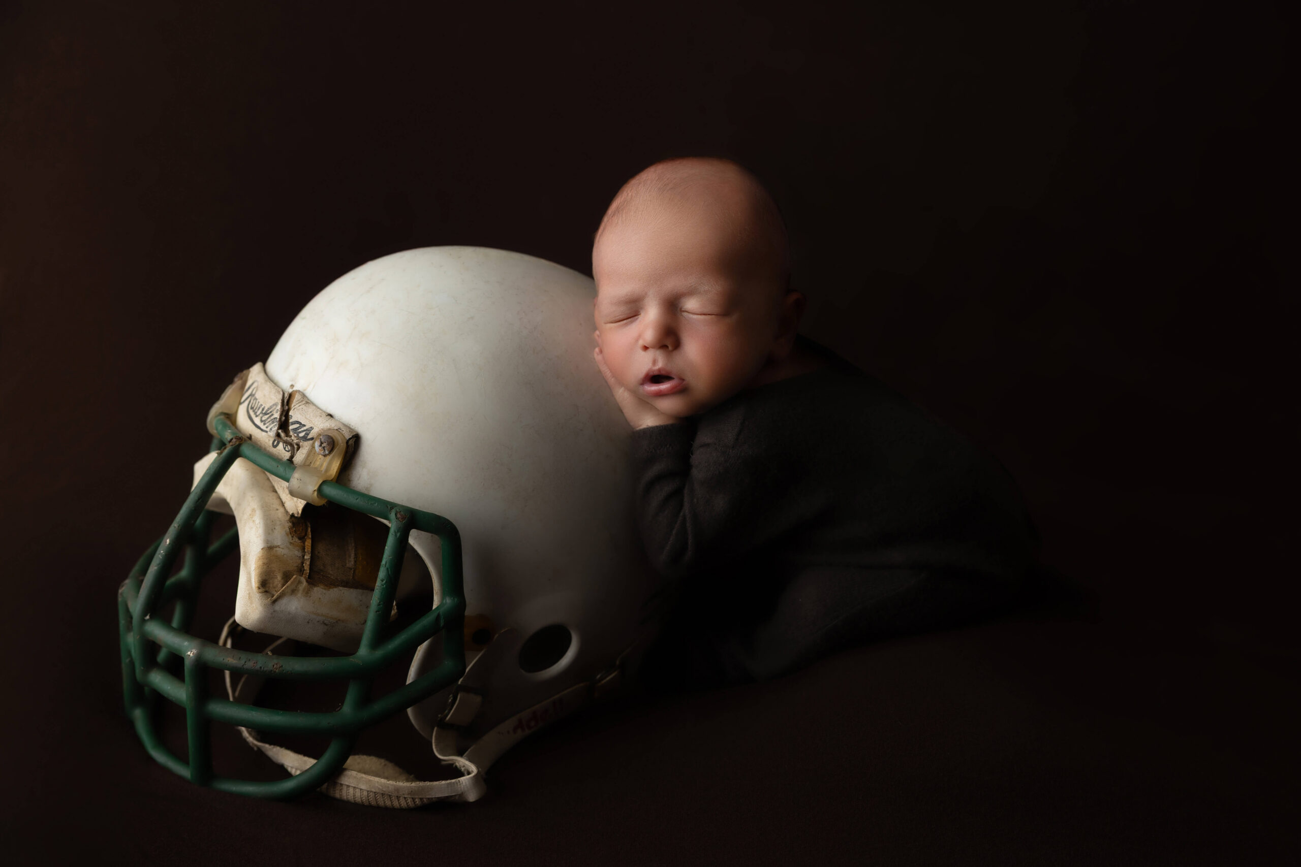 A sleeping newborn resting on a football helmet in a black onesie