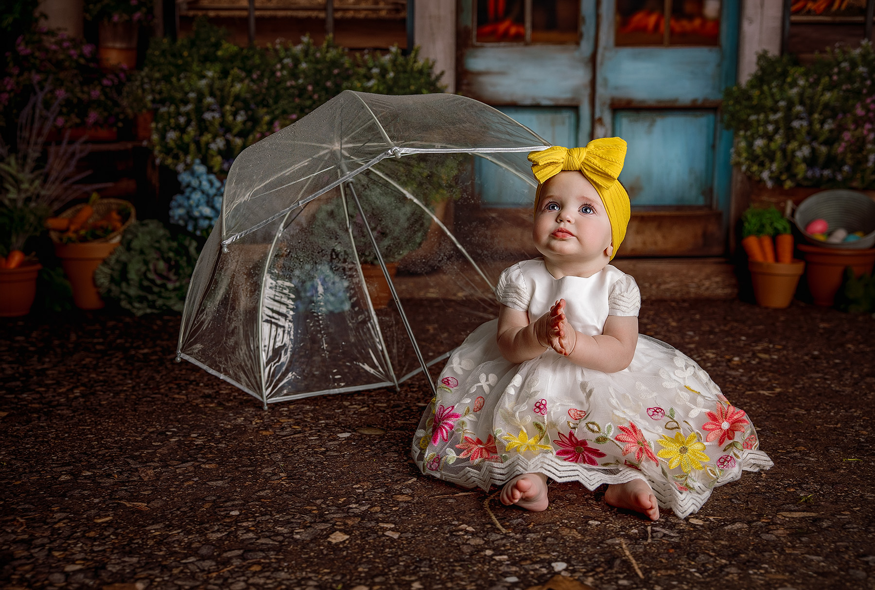 A clapping baby girl in a white and flower covered dress sits next to a clear umbrella looking up