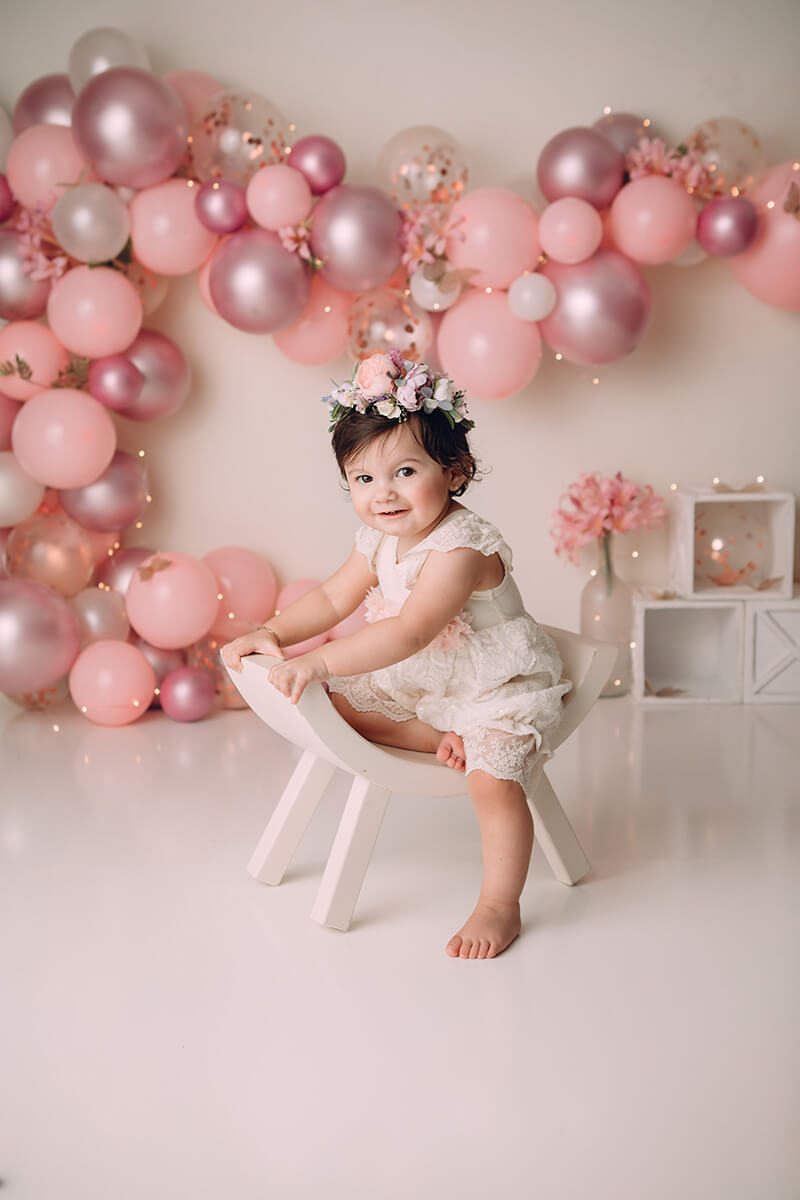 A smiling baby girl in a white lace dress sits in a curved white small chair