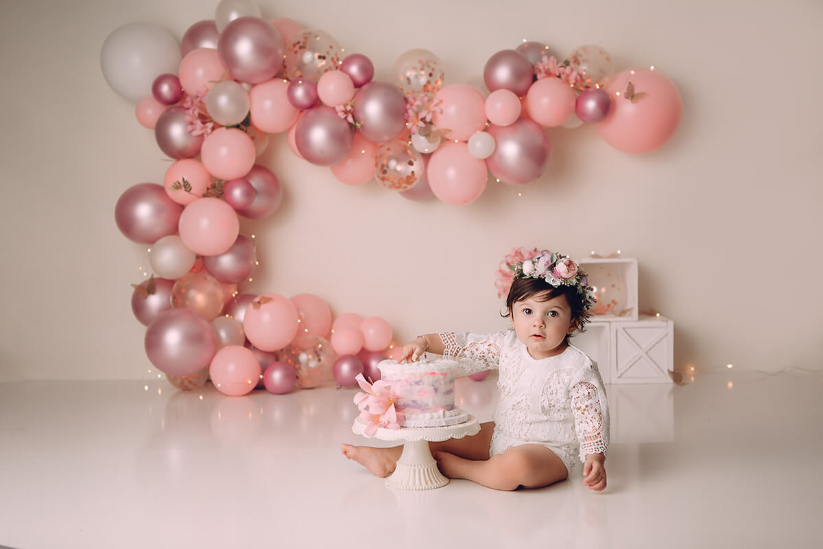 A young baby girl in a lace onesie sits in a studio shoving her hand into a pink cake after visiting toy stores in Peoria, IL