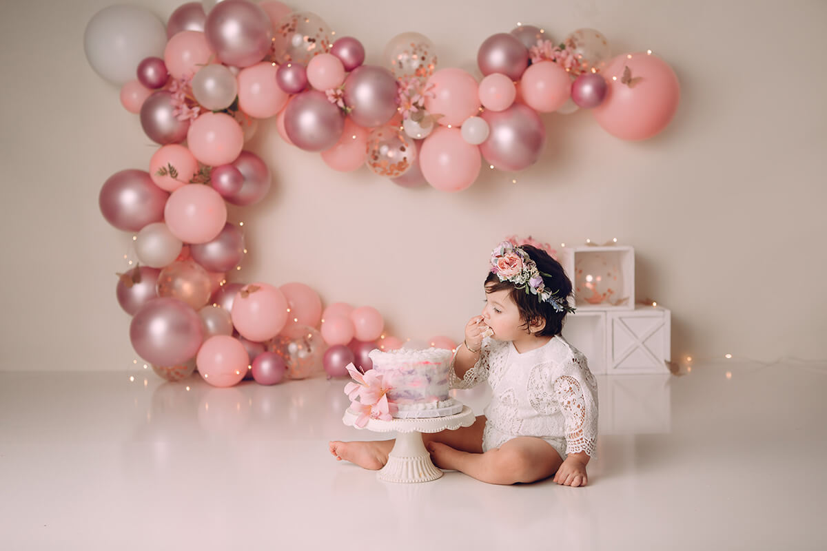 A toddler girl in a white lace outfit sits in a studio eating a cake for her birthday after visiting toy stores in Peoria, IL