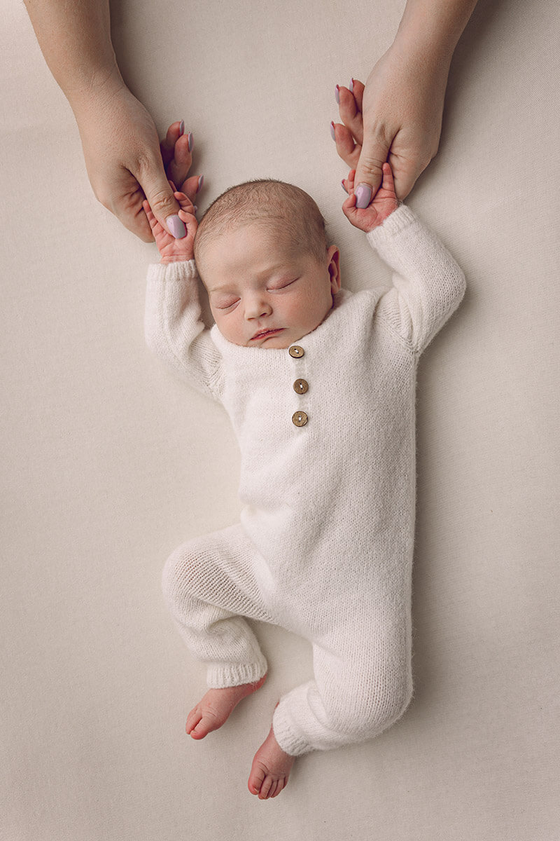 A sleeping newborn in a white onesie lays while holding mom's thumbs above its head