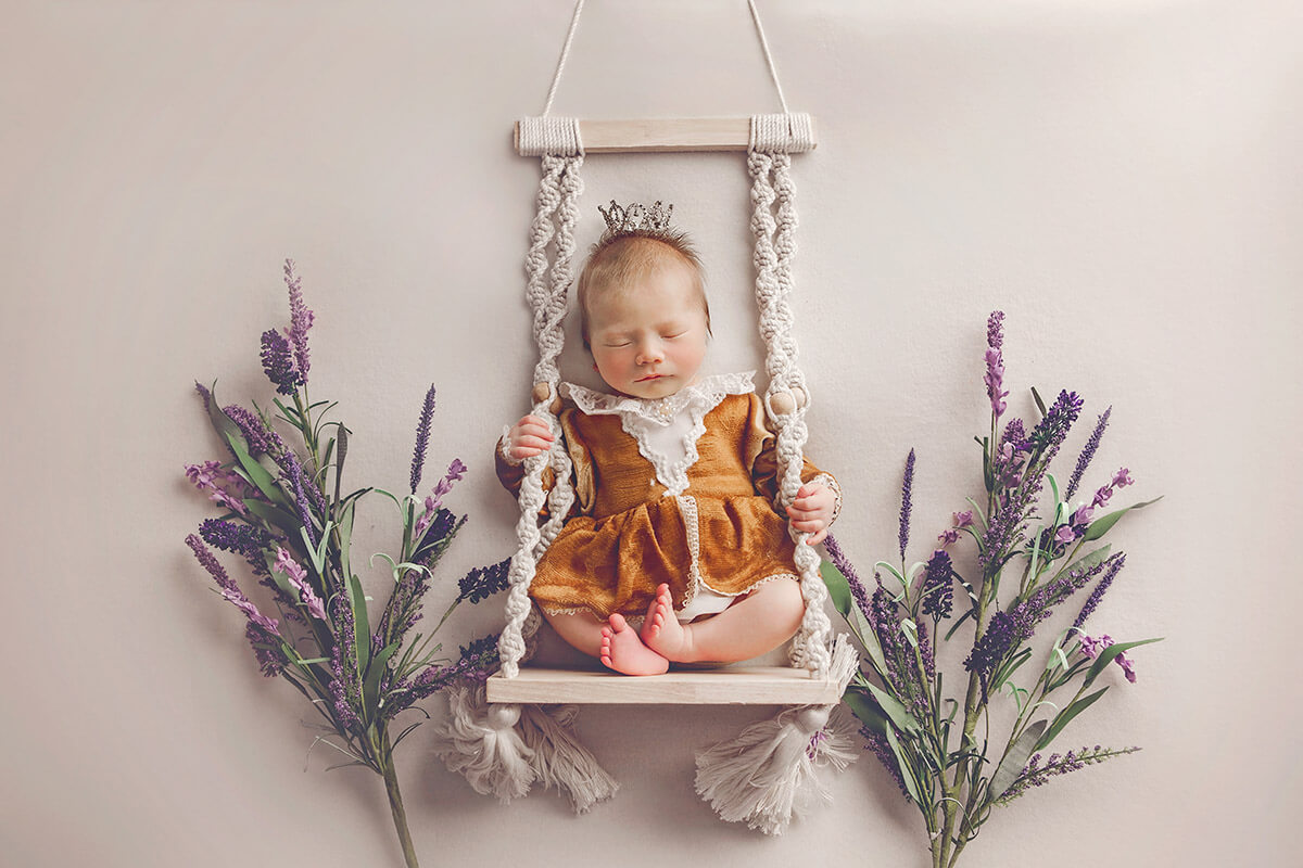 A newborn baby sleeps while sitting on a wood and rope swing in a gold dress surrounded by lavender after visiting baby boutiques in Champaign, IL