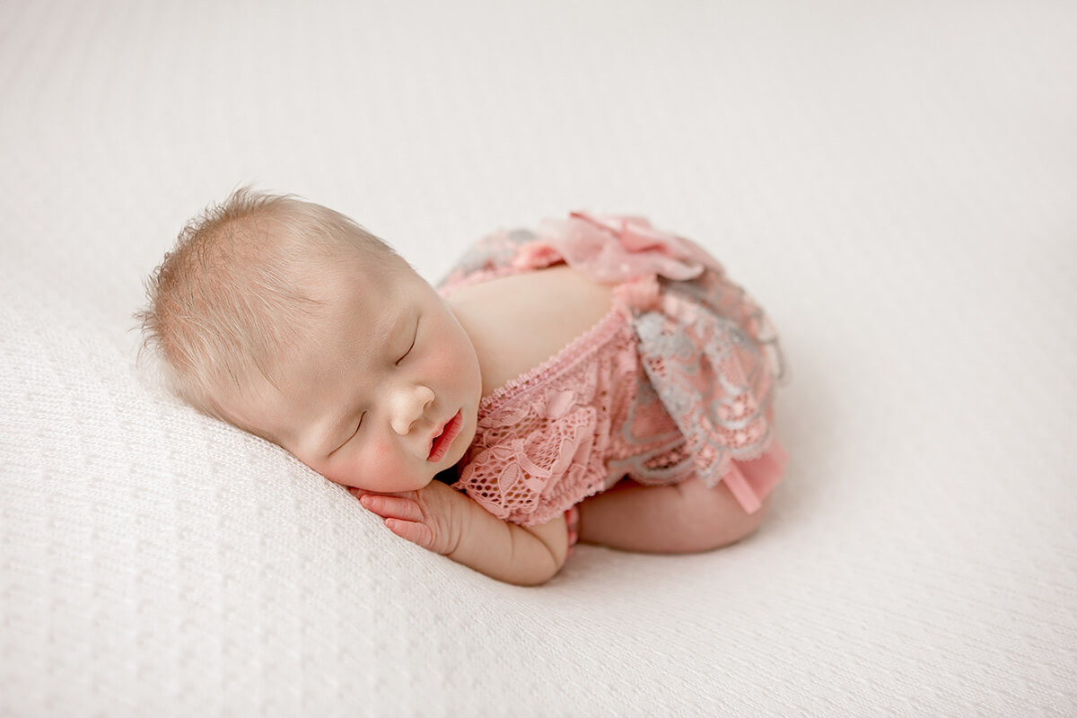 A sleeping newborn baby girl in froggy pose in a pink lace onesie on a white bed after visiting baby boutiques in Champaign, IL