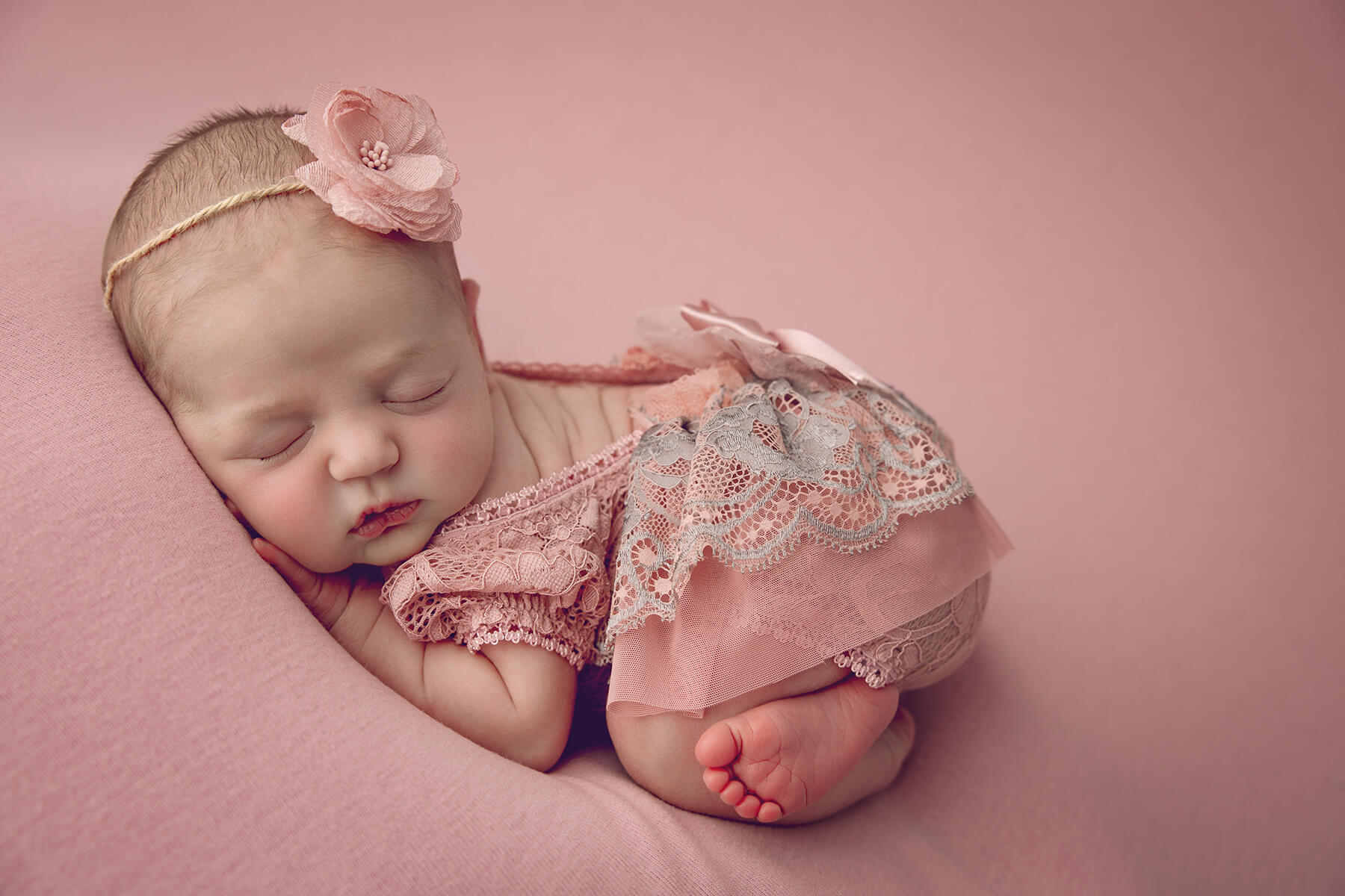 A sleeping newborn in a pink lace onesie on a pink bed in a studio