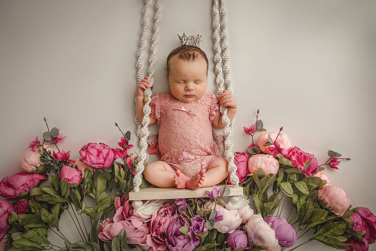 A newborn baby sits on a wooden swing above an array of pink flowers after mom enjoyed birthing classes in Champaign, IL