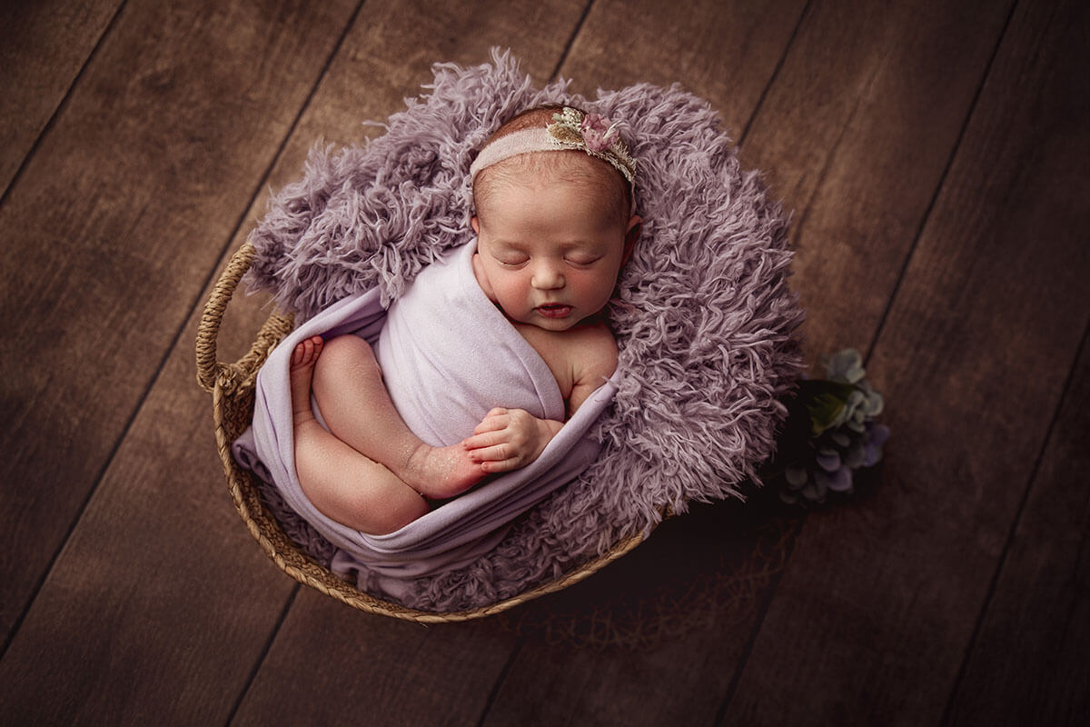 A newborn baby girl in a pink swaddle in a woven basket on wood floor