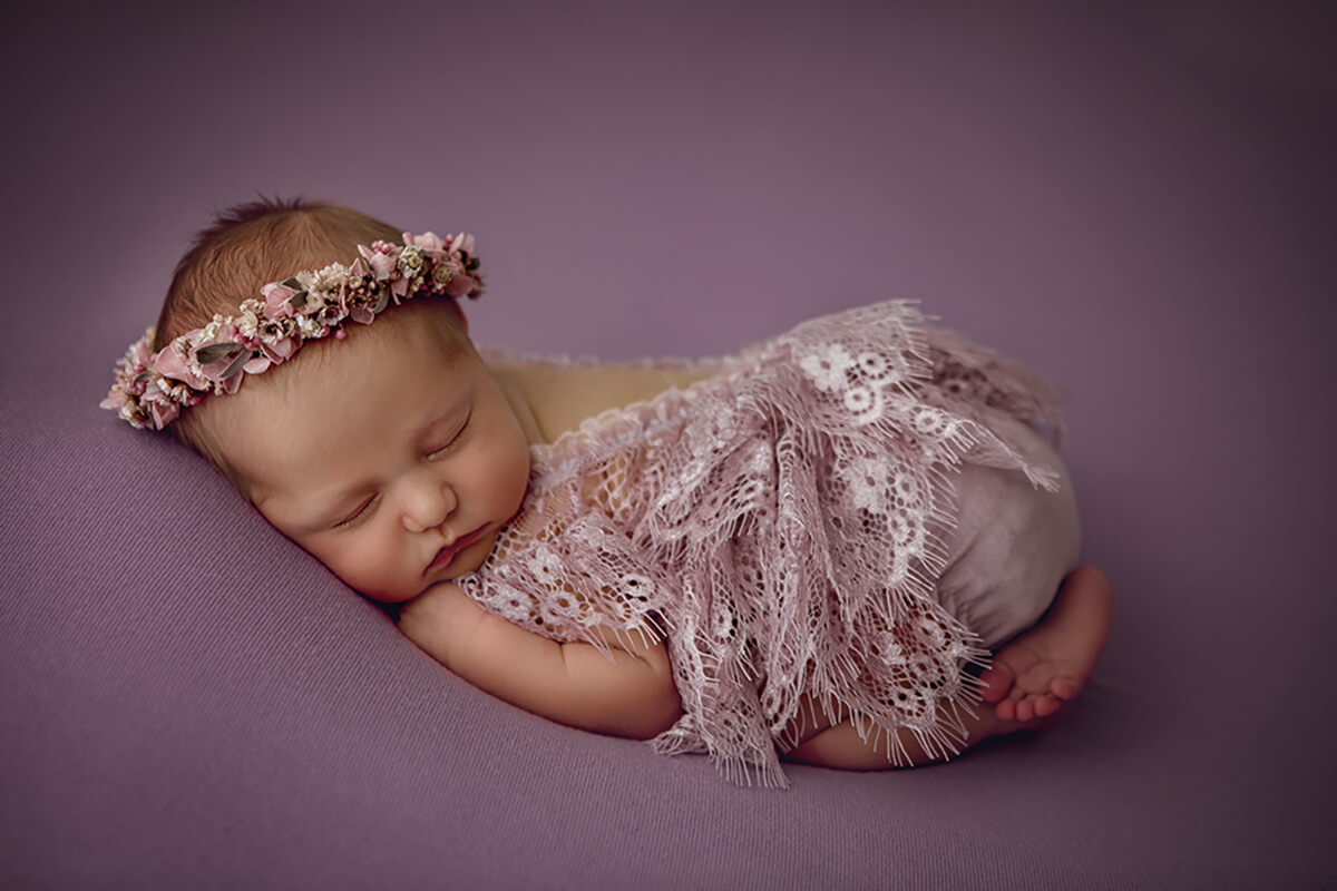 A sleeping newborn baby girl in froggy pose in a pink lace dress and matching flower headband after meeting lactation consultants in Champaign, IL
