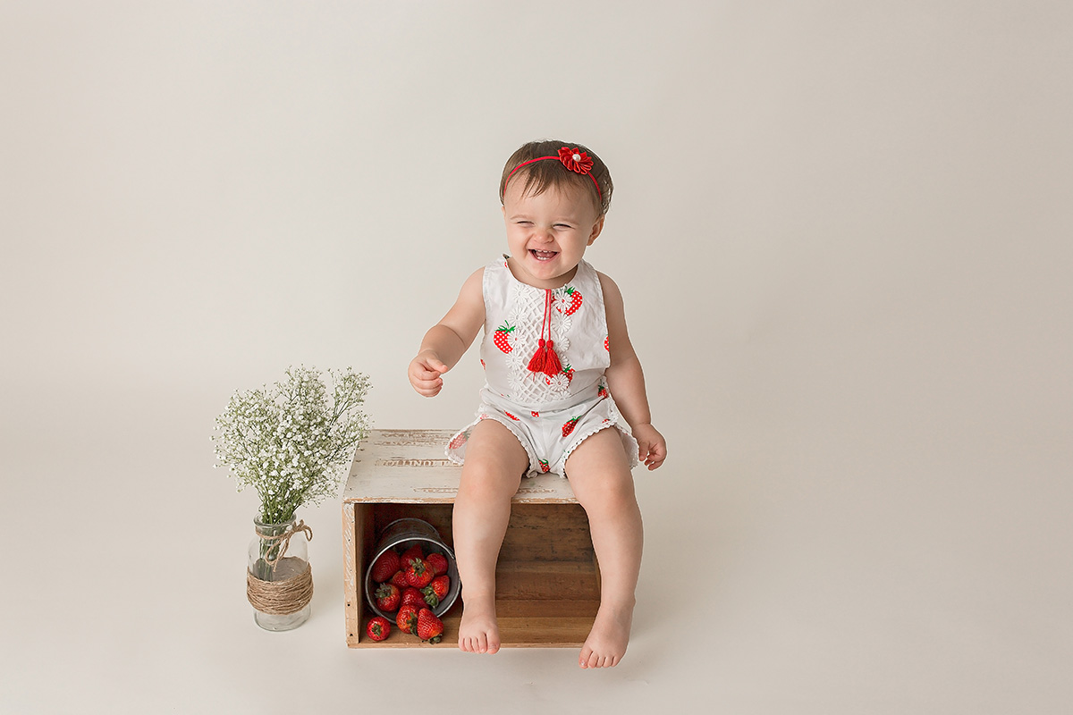A giggling toddler girl in a strawberry jumper sits on an apple box with a bucket of strawberries after enjoying montessori schools in springfield il