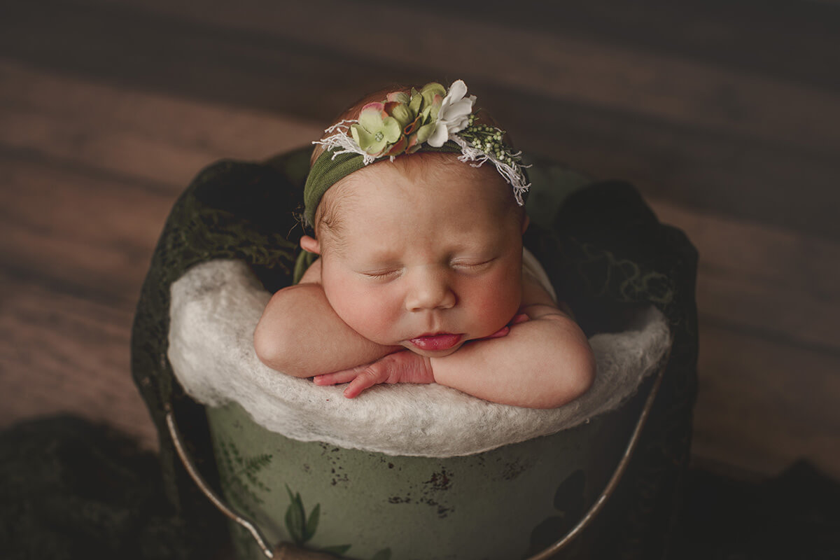 A newborn baby sleeps resting on the edge of a metal bucket with a flower headband after meeting postpartum doulas in Champaign, IL