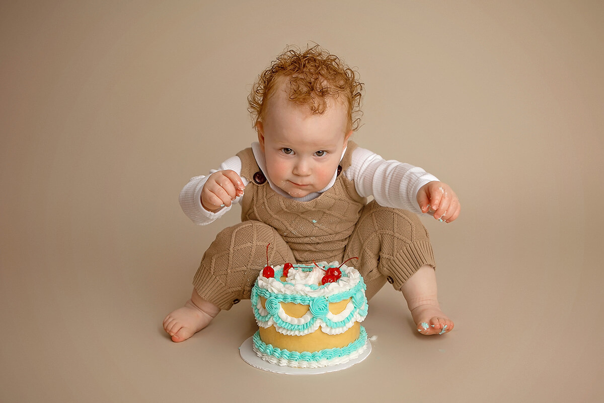 A young boy in tan overalls sits behind a cherry topped cake in a studio