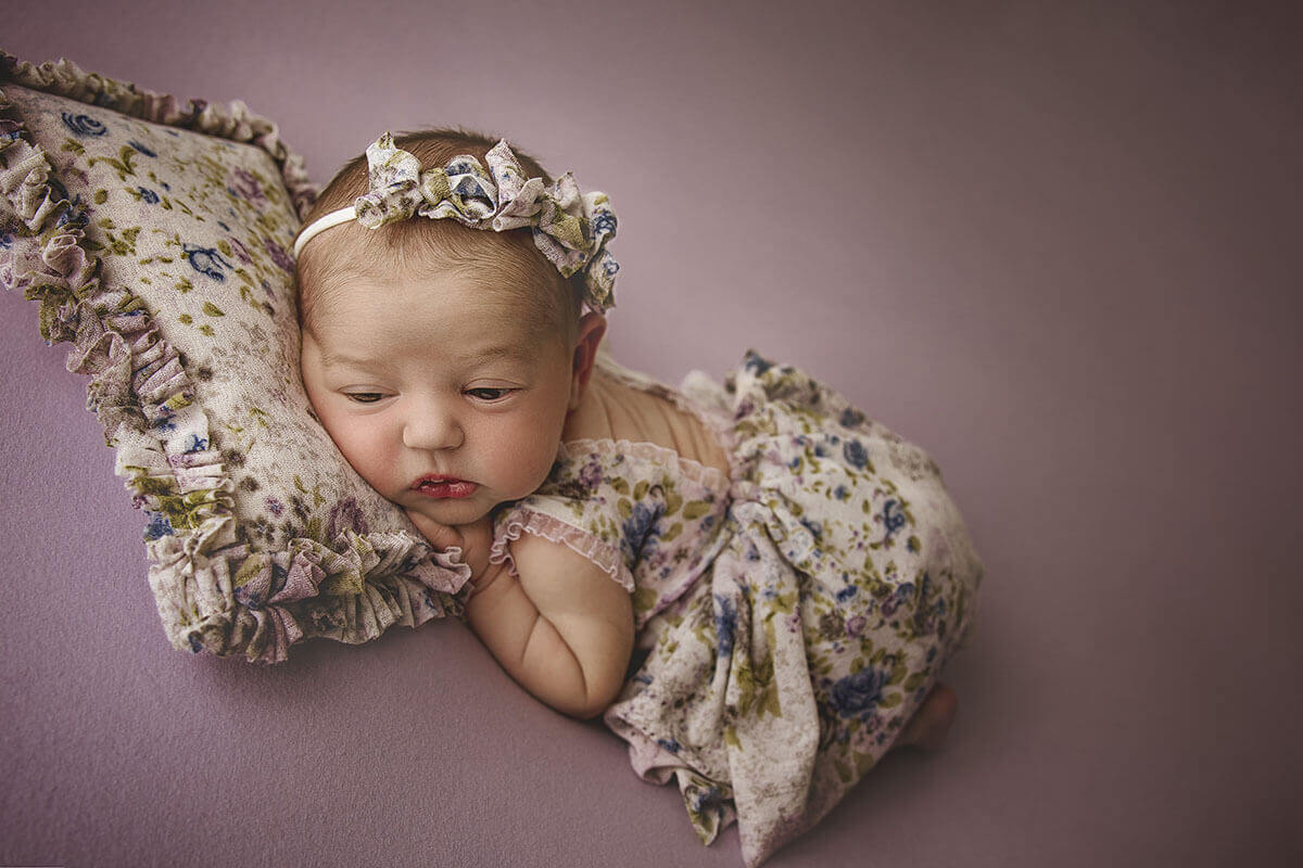 A newborn baby girl in a floral print dress lays with eyes open in froggy pose