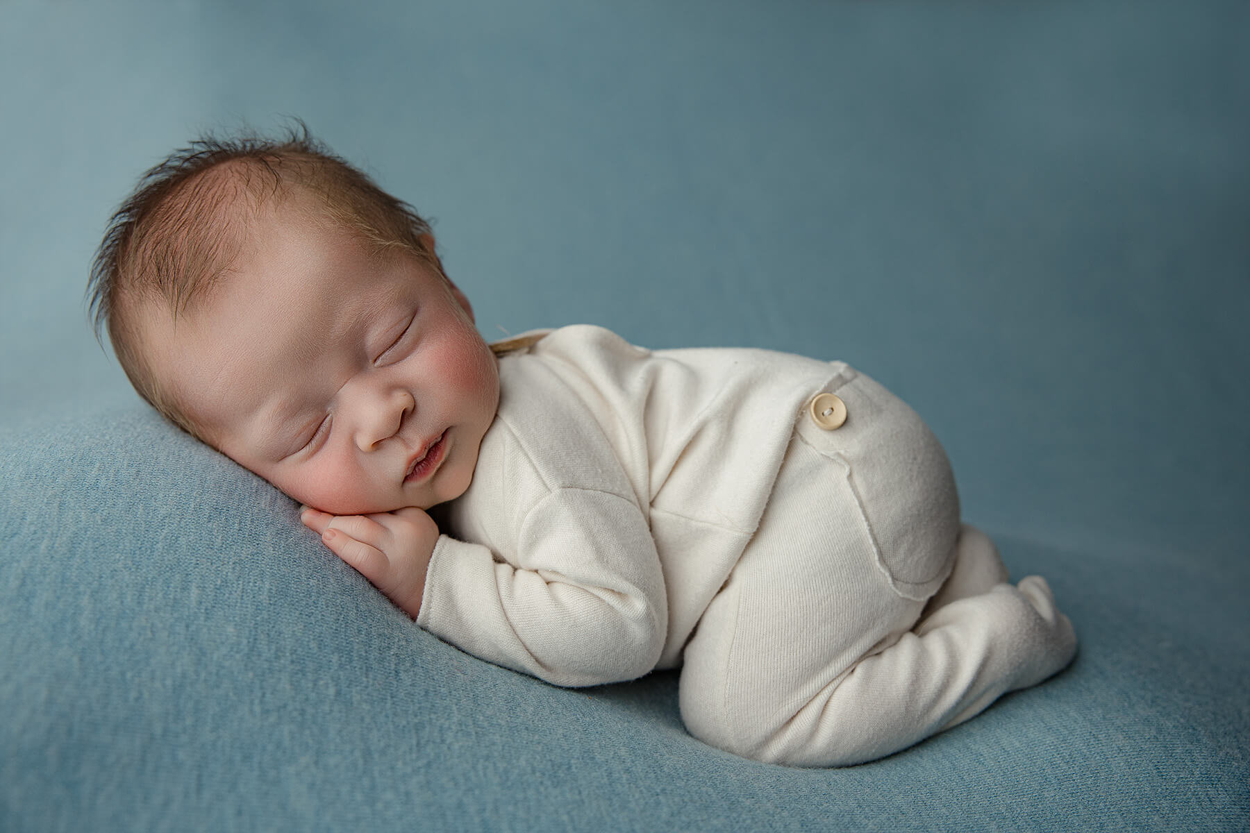 A sleeping newborn in froggy pose wearing a white onesie on a blue bed