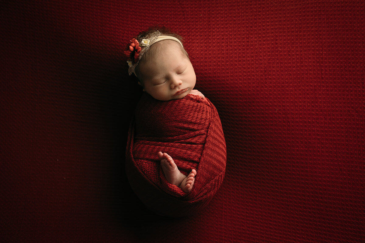 An infant in a red swaddle sleeping with toes out and a red flower headband
