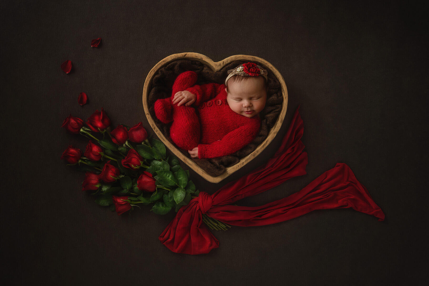 A sleeping newborn girl in a red onesie in a wooden bowl framed by red roses thanks to obgyns in champaign il