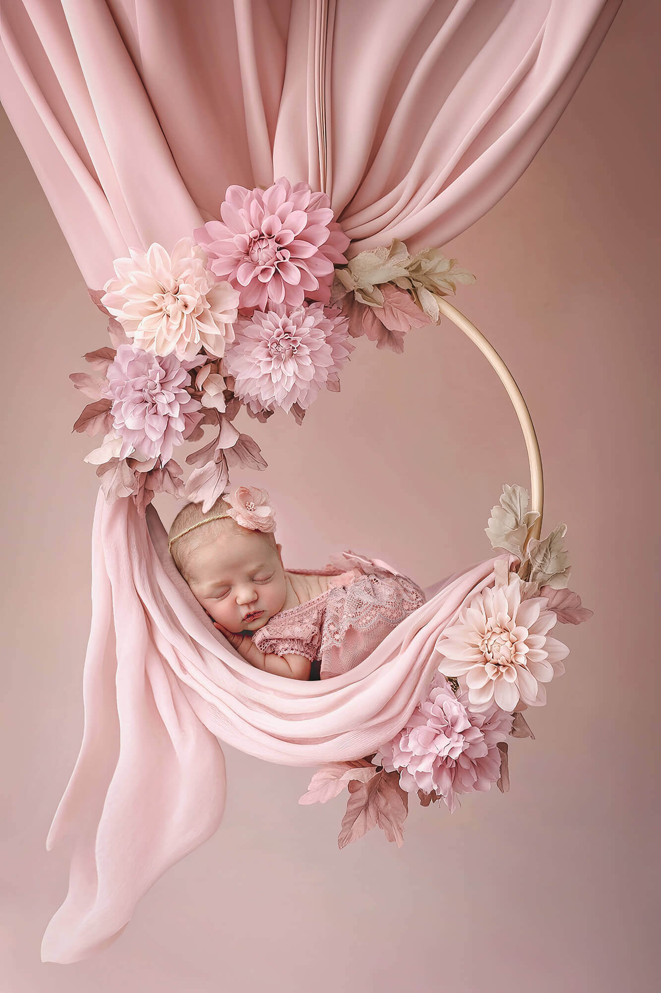 A sleeping newborn girl in a pink lace dress on a hoop swing in froggy pose held by flowers and pink drape