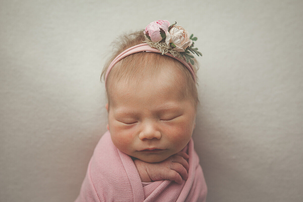 A baby girl sleeps in a pink swaddle with a hand poking out and a matching pink flower headband after meeting pediatric dentists in champaign il