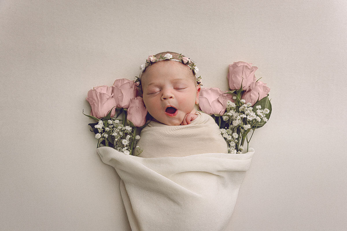 A yawning baby girl with a flower headband in a white swaddle fulled with pink roses