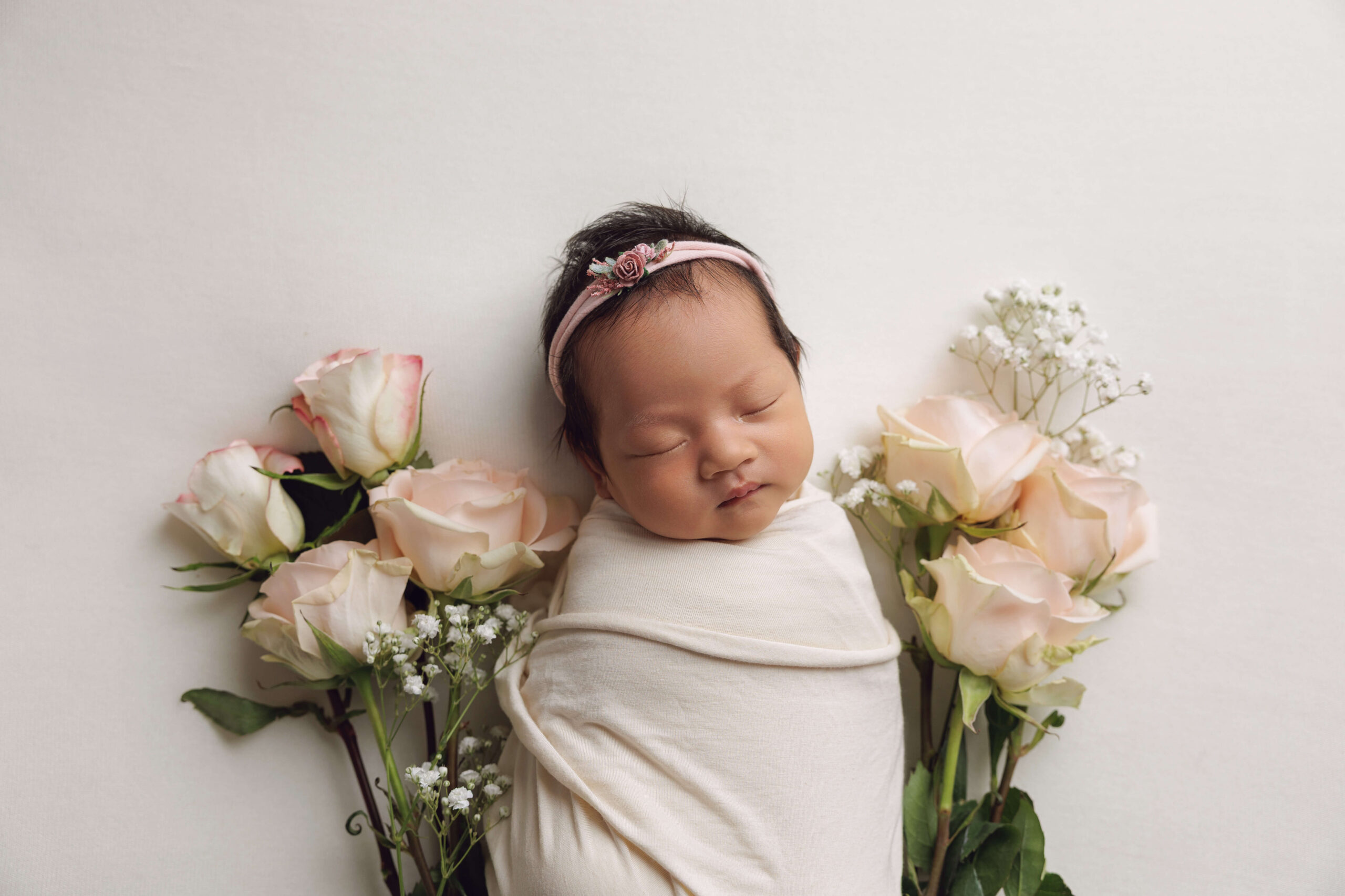 A sleeping newborn in a white swaddle surrounded by pink roses with a matching pink headband after meeting pediatricians in champaign il