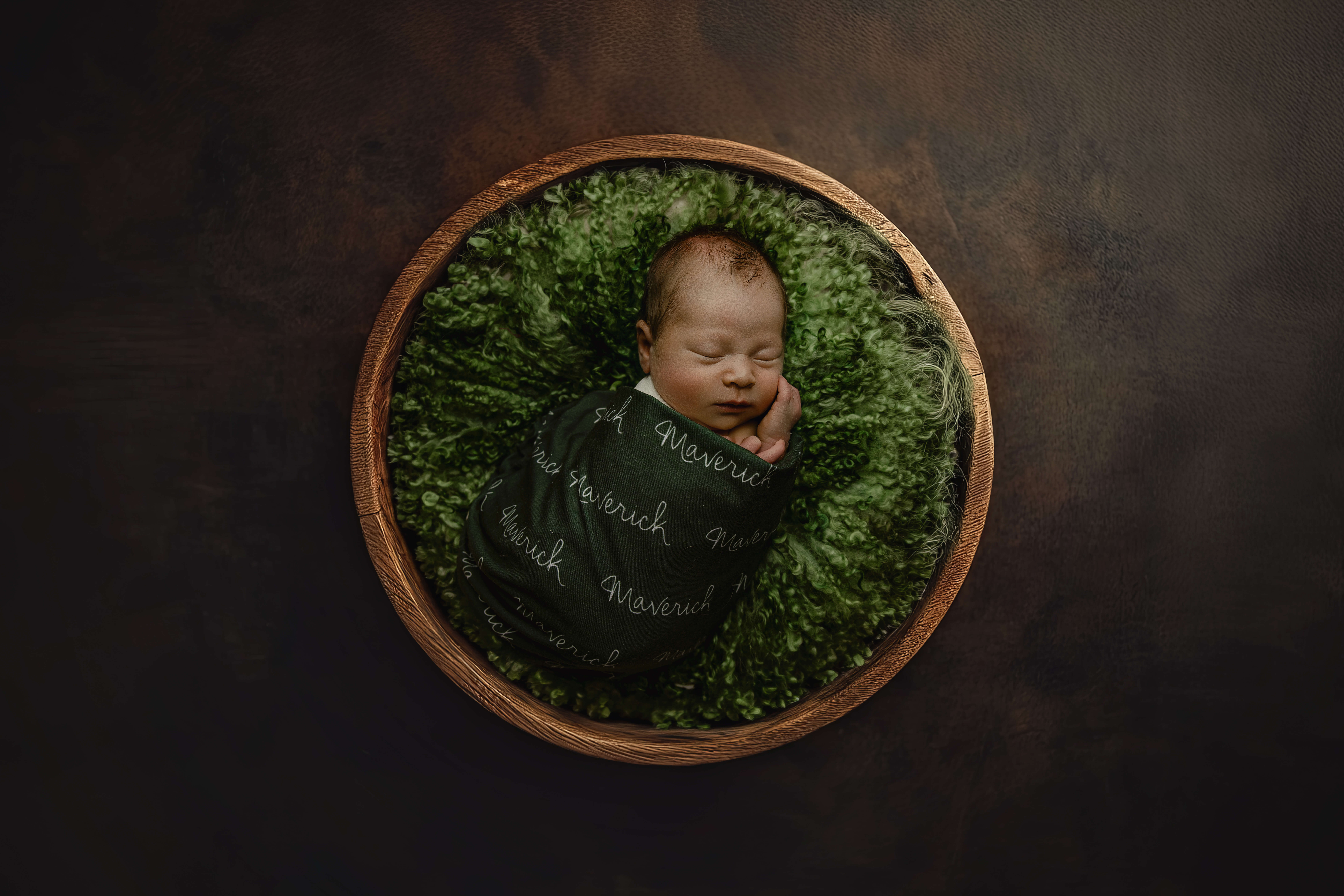 A newborn baby sleeps wrapped in a custom green blanket in a wooden bowl after mom enjoyed a prenatal massage in champaign, IL