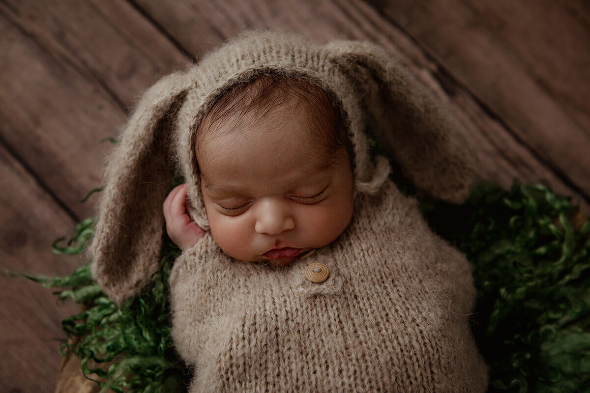 A newborn baby sleeps in a tan knit bunny onesie on wood floor after mom benefitted from a prenatal massage in champaign, IL