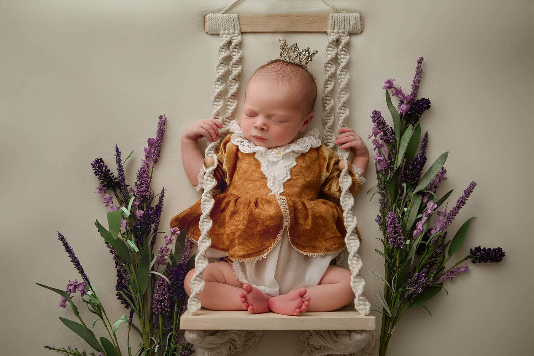 A baby girl in an orange dress sleeps on a macrame swing with lavender and a crown