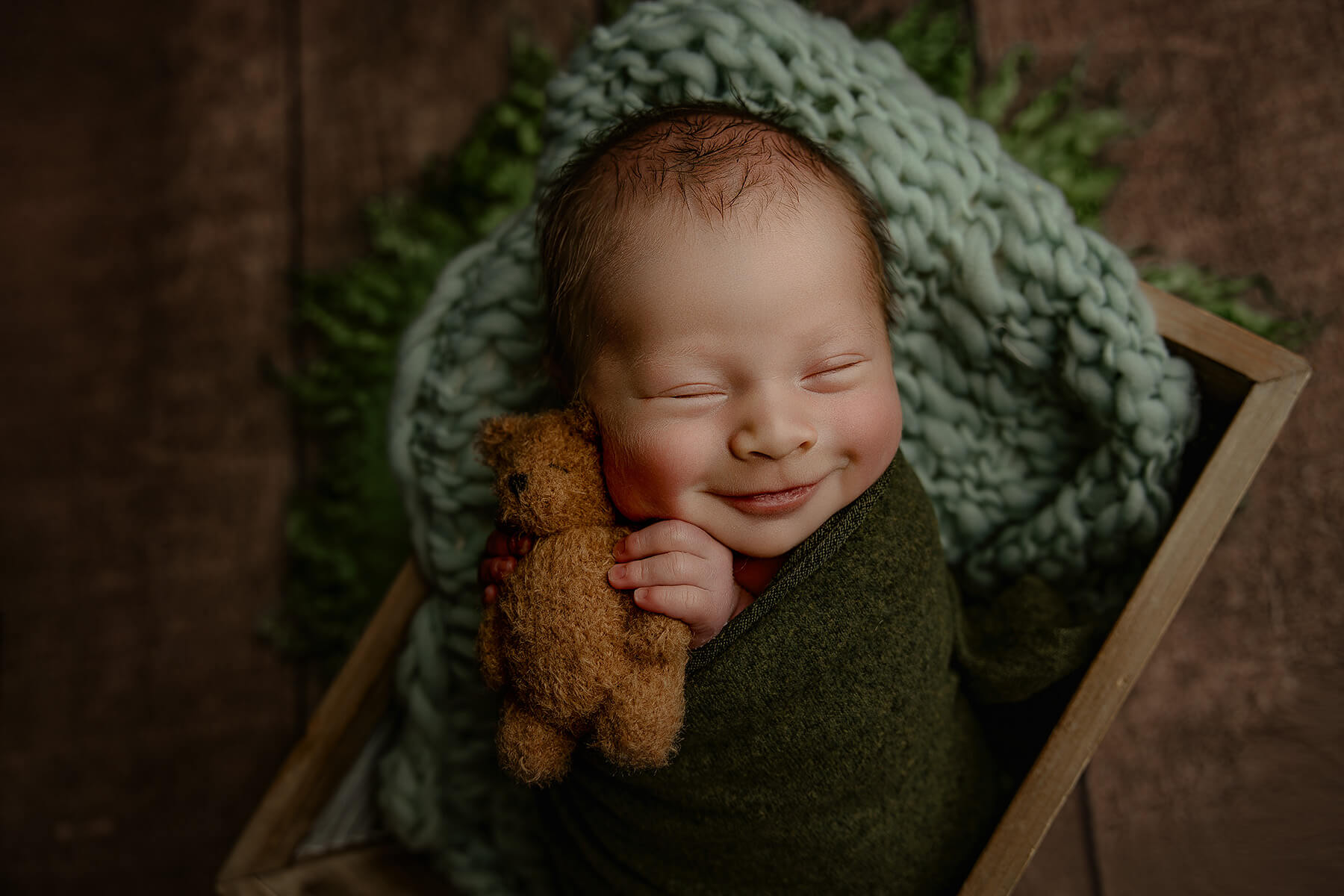 A smiling newborn sleeping with a brown teddy in a wooden box