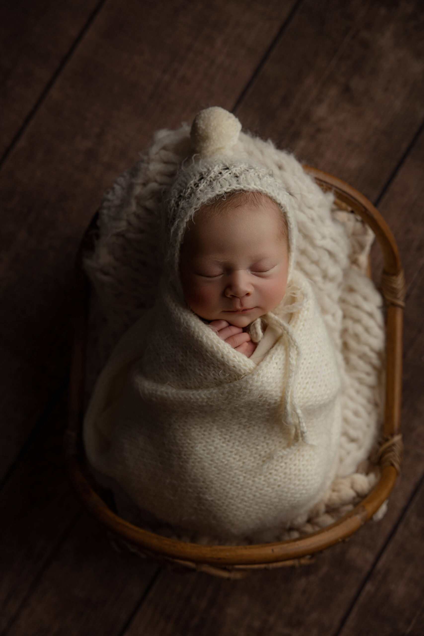 A sleeping newborn baby in a white knit swaddle and matching sleep cap in a wicker basket after using here together birth